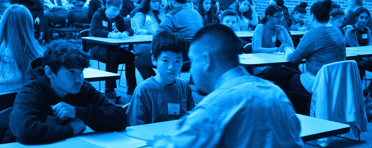 The image depicts a crowded classroom setting, with numerous students seated at desks and engaged in various activities. The lighting in the room appears to be predominantly blue, creating a distinctive atmosphere.