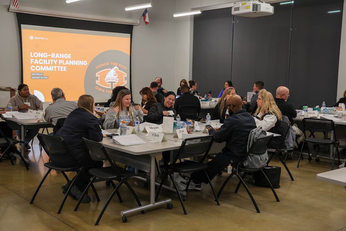 Committee members gather around tables with a presentation screen visible in the background.