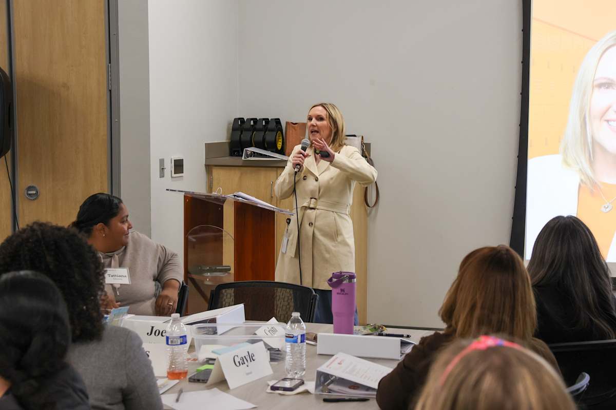 A woman in a beige coat stands at a podium addressing people seated around a table.