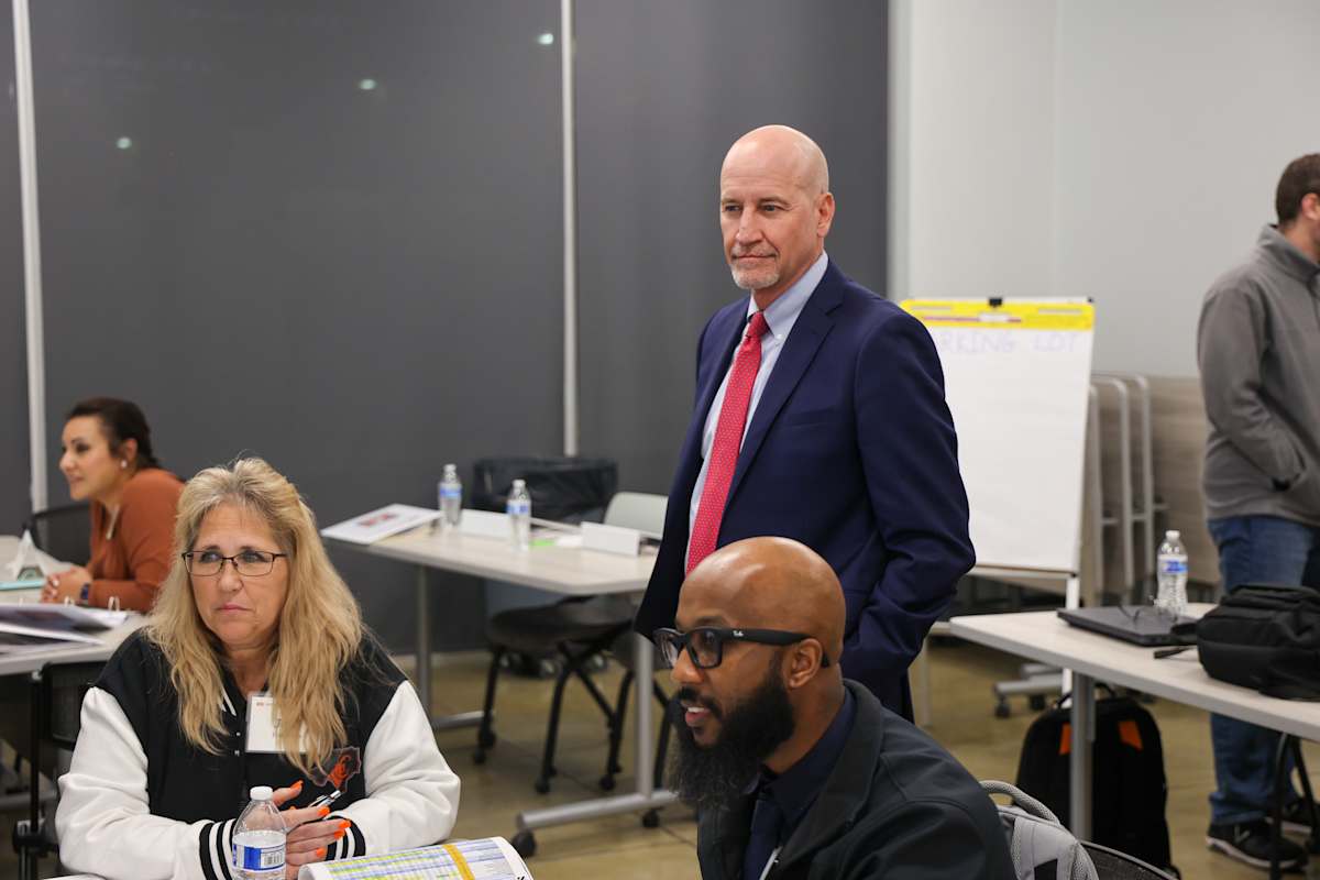A man in a suit stands while participants sit at tables during a facilities planning meeting.