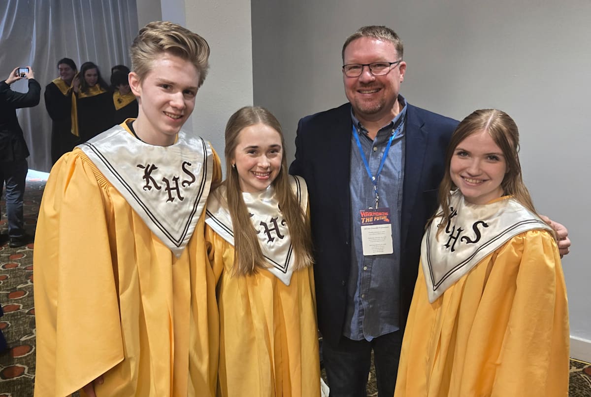 Three students in yellow robes with stoles that say KHS pose together with a man in a suit jacket and a lanyard around his neck.