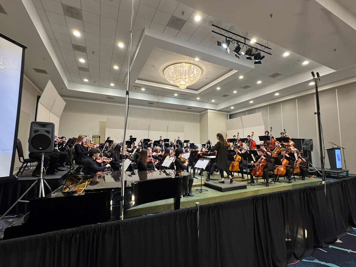 A woman stands in front of a large group of students with instruments and music stands on a stage.