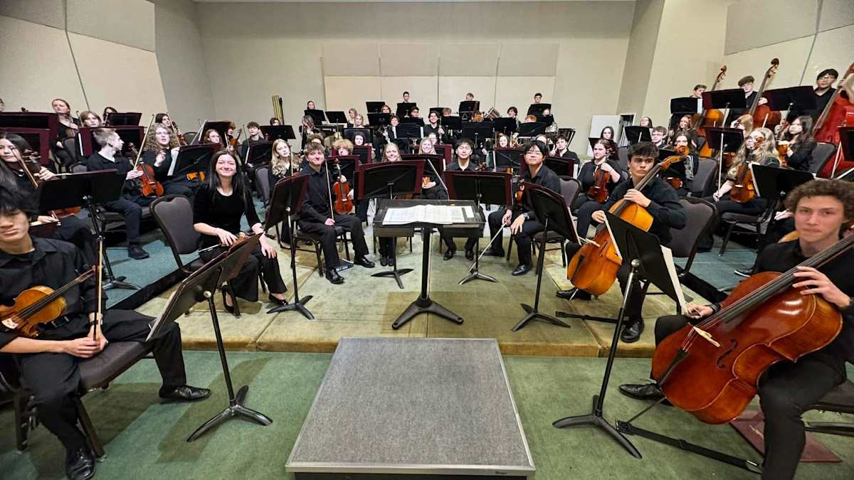 An orchestra of students dressed in black with instruments sit in a half circle and smile.