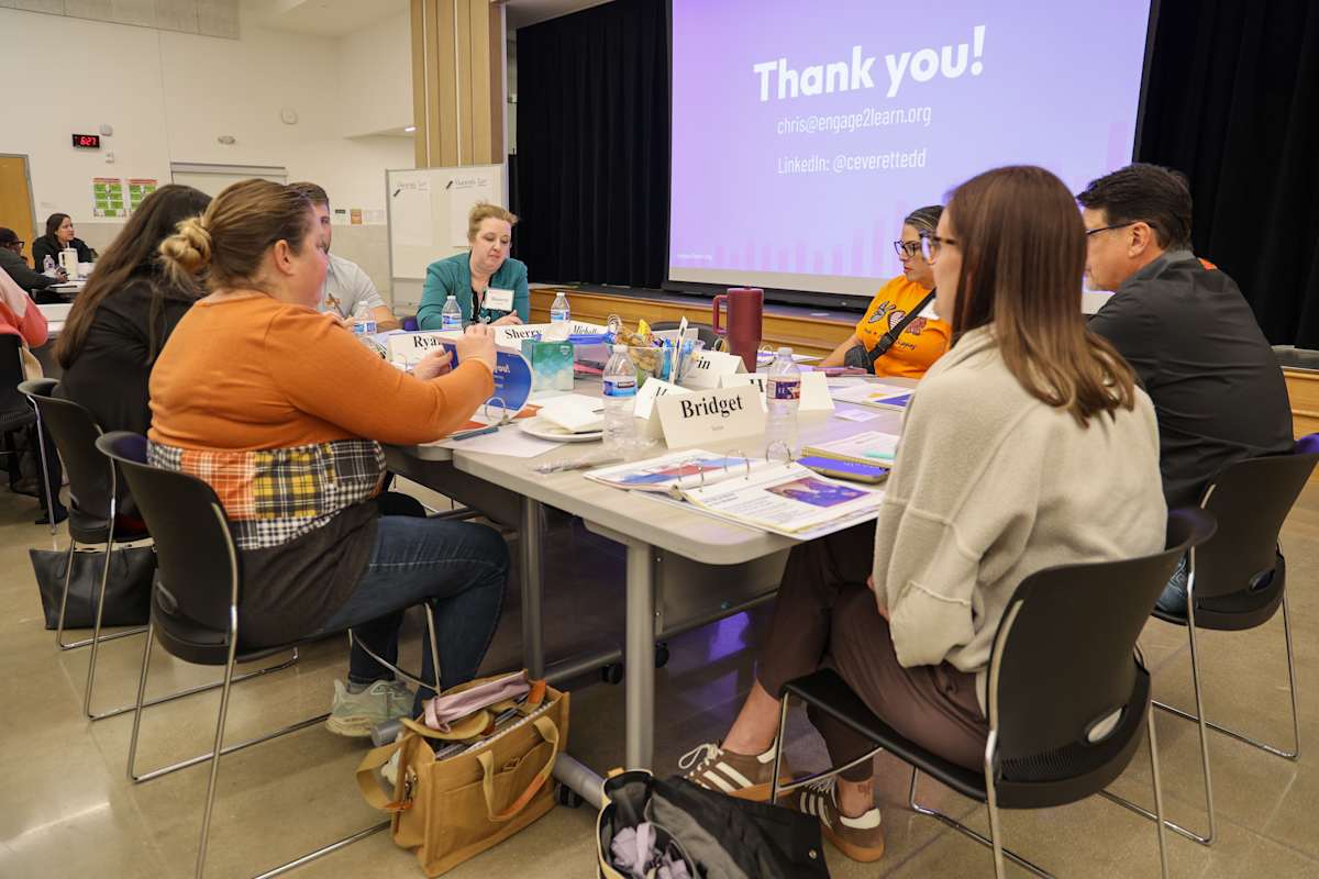 A group gathers around a table for discussion while a Thank you message is projected behind them.