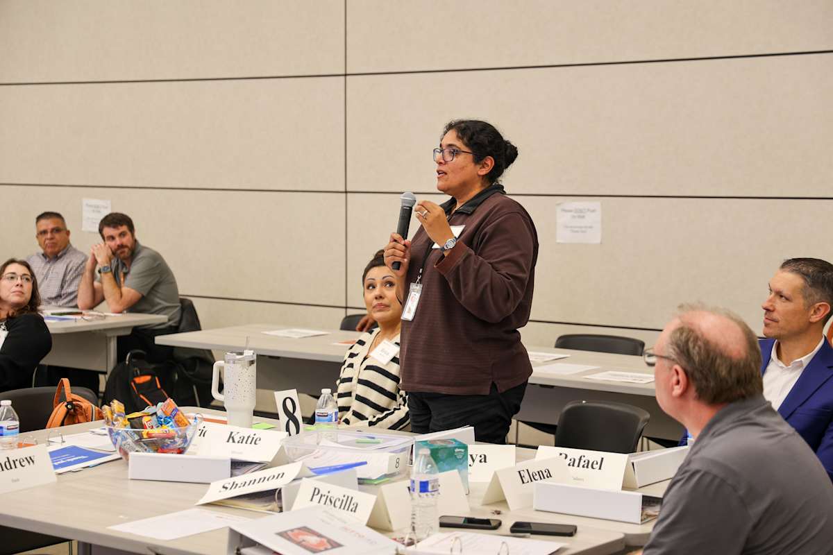 A participant speaks into a microphone while others sit around a meeting table.