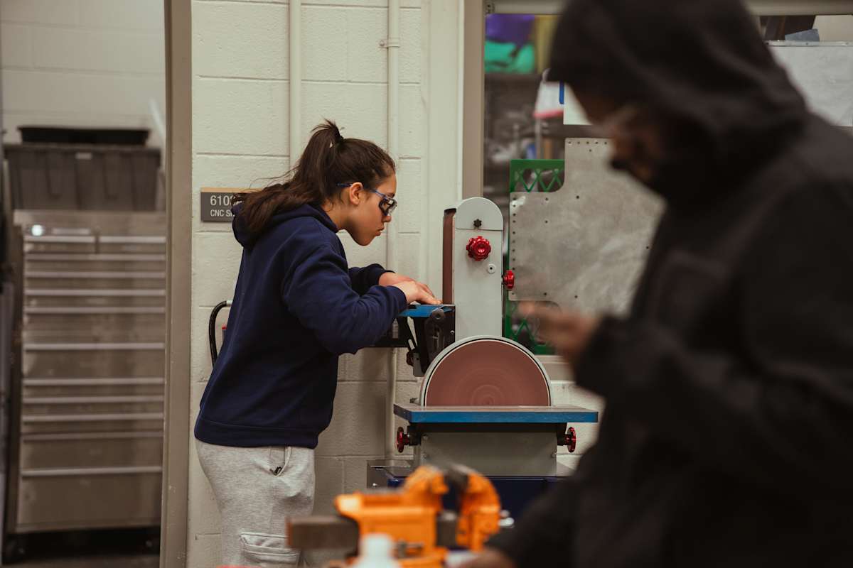 A student in the foreground uses a sanding machine in a workshop