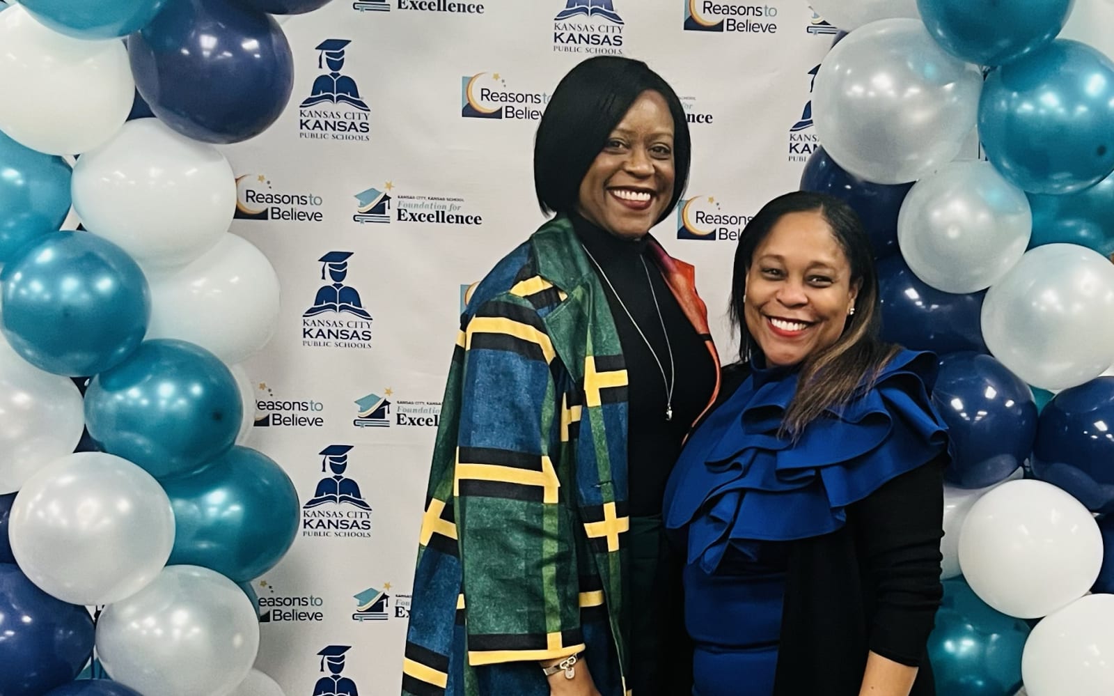 Two smiling women wearing blue and green stand in front of balloons and a KCKPS background.
