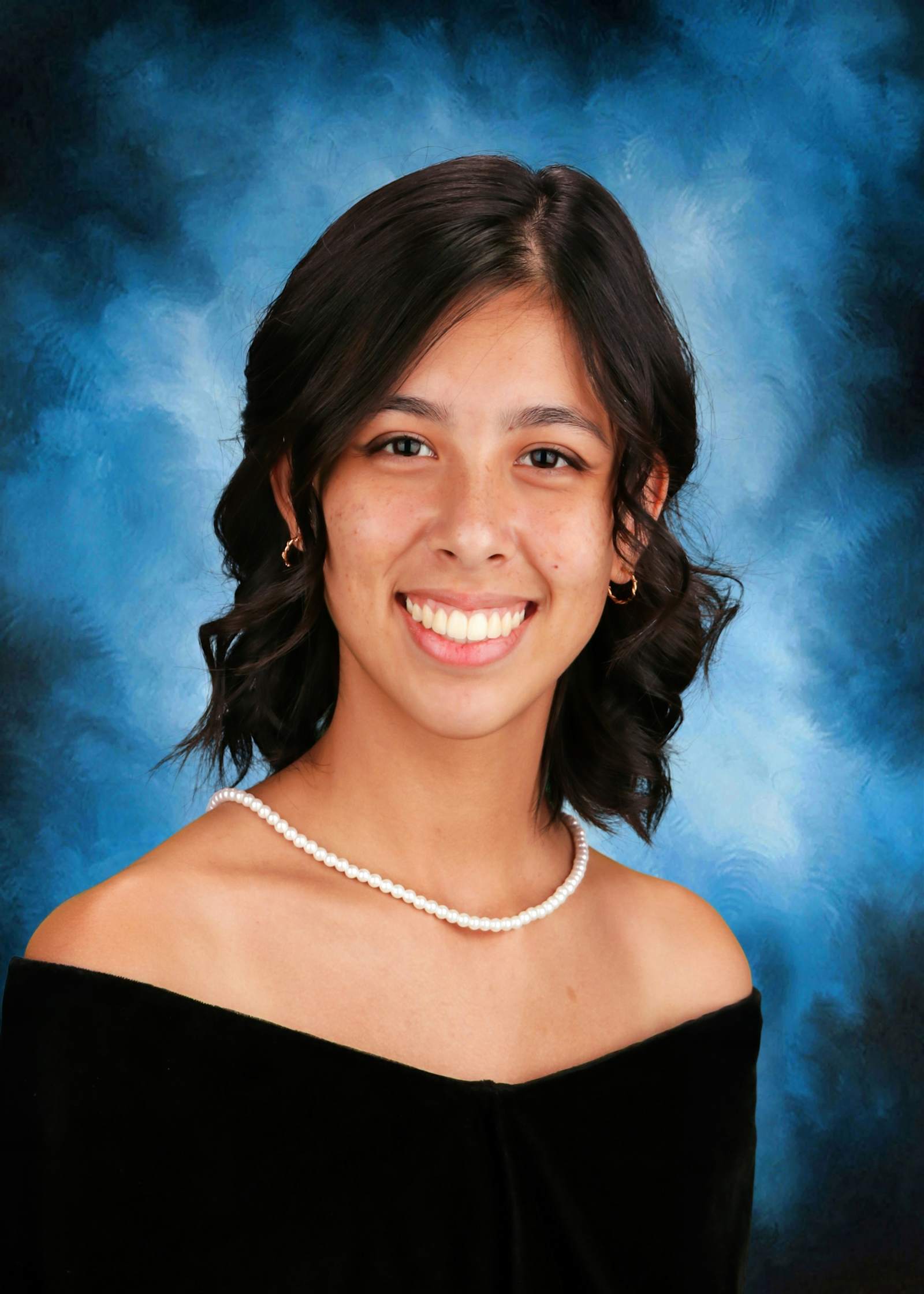 A young woman with dark hair and a warm smile, wearing a black off-the-shoulder top, stands against a vibrant blue background with a soft, hazy effect.