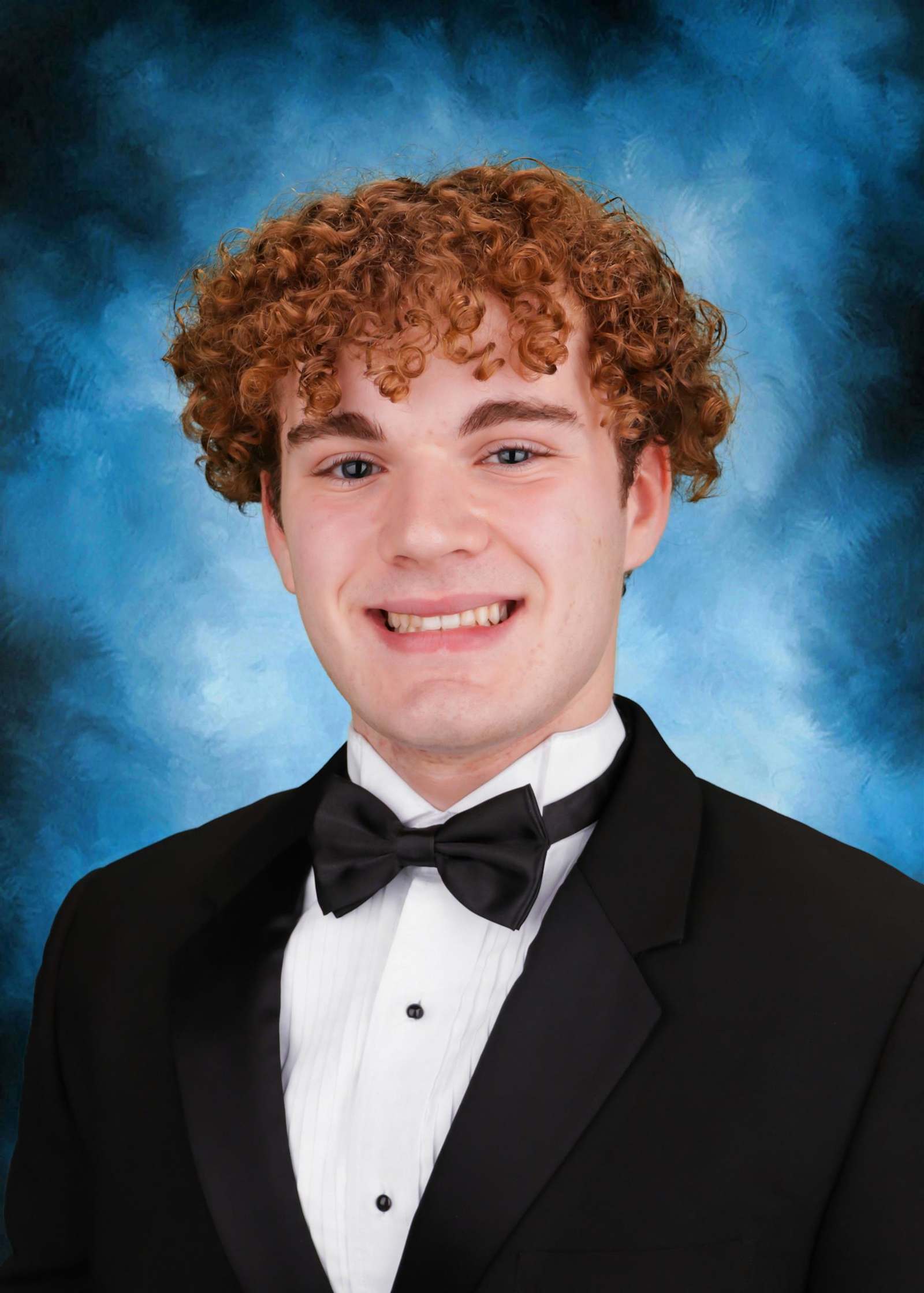 The image shows a young man with curly red hair wearing a black tuxedo and bow tie, smiling against a blue smoky background.