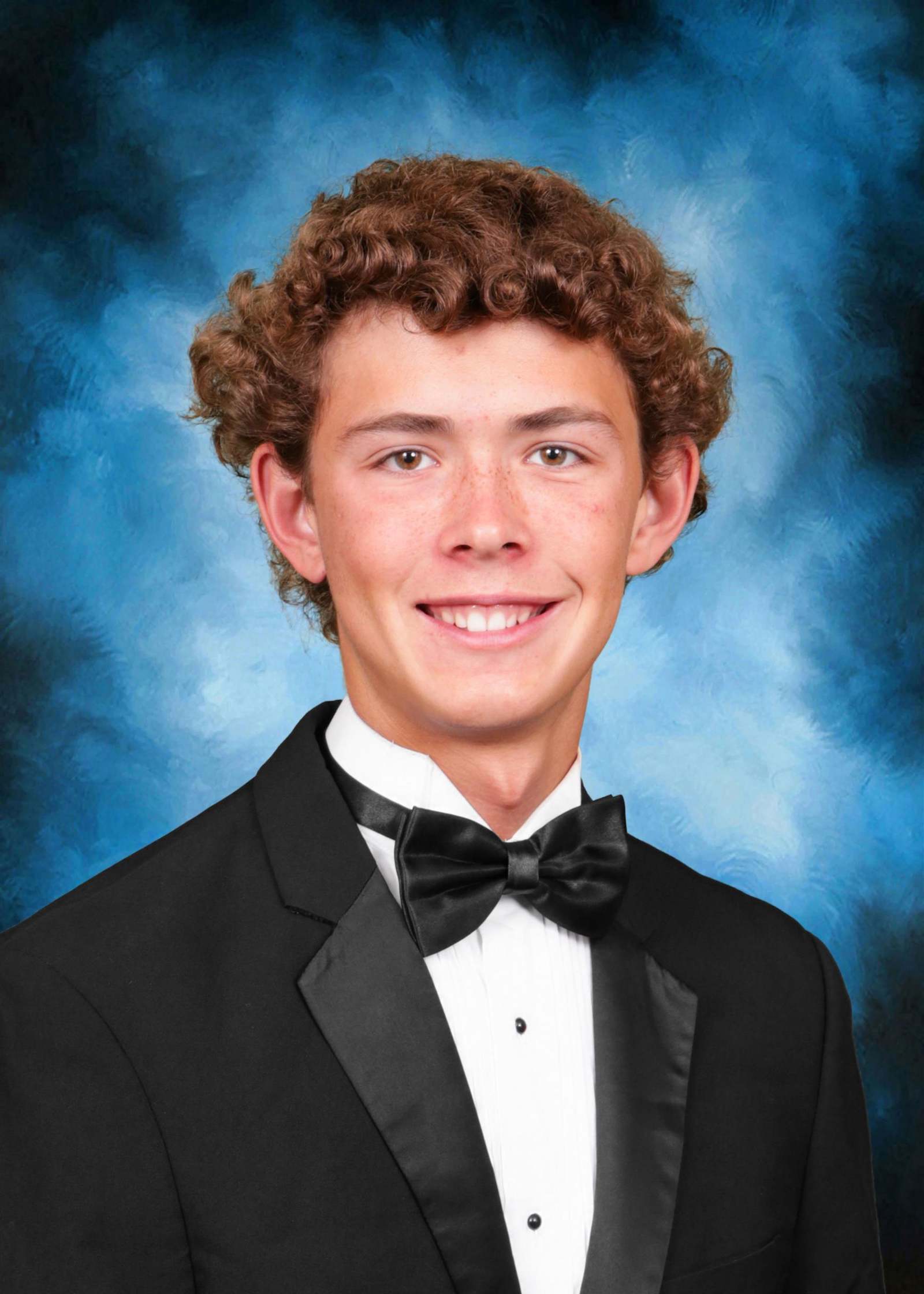 The image shows a young man with curly brown hair wearing a black tuxedo and bow tie, smiling against a blue and smoky background.