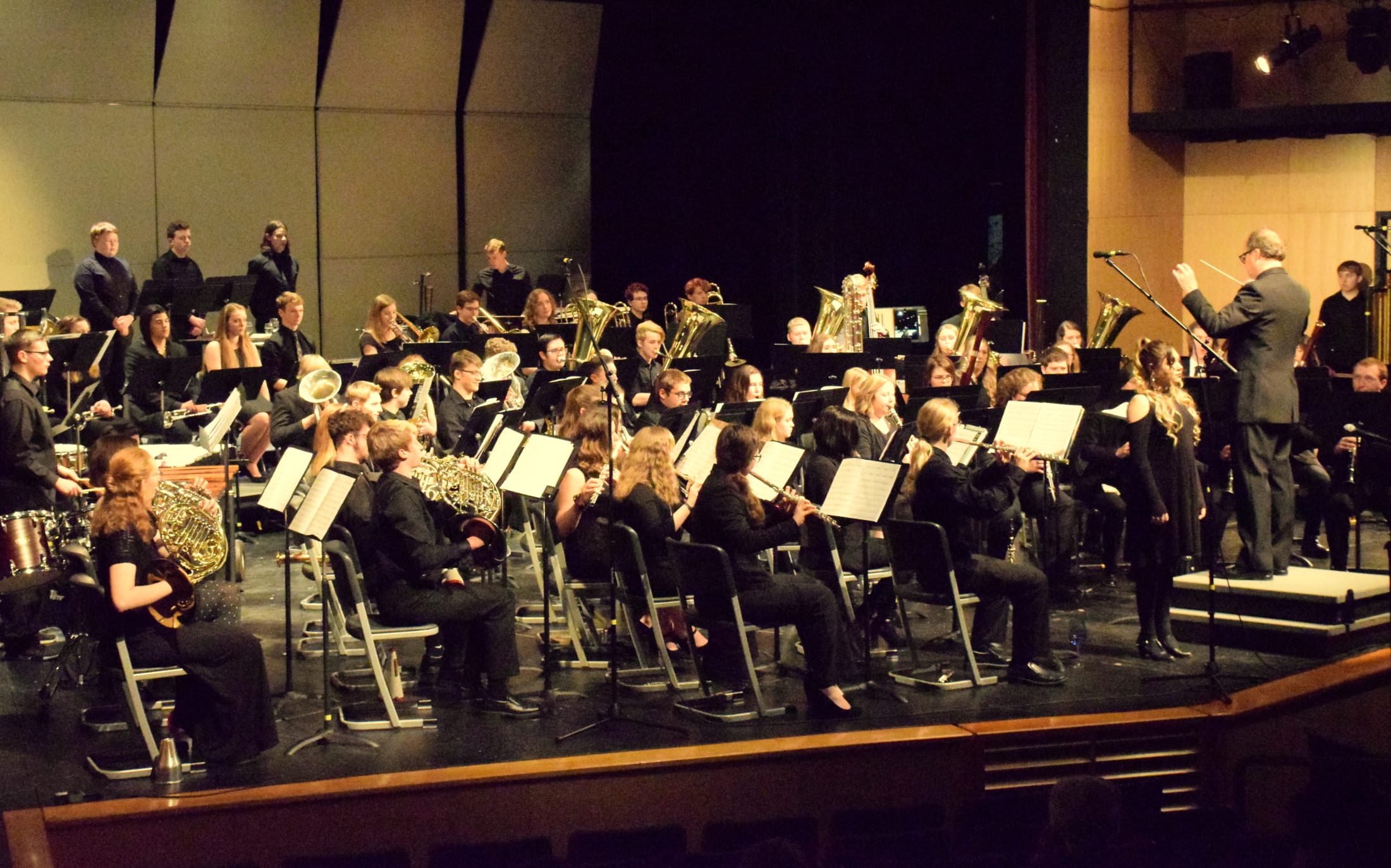A large orchestra performing on stage, with musicians playing various instruments and the conductor leading the ensemble.