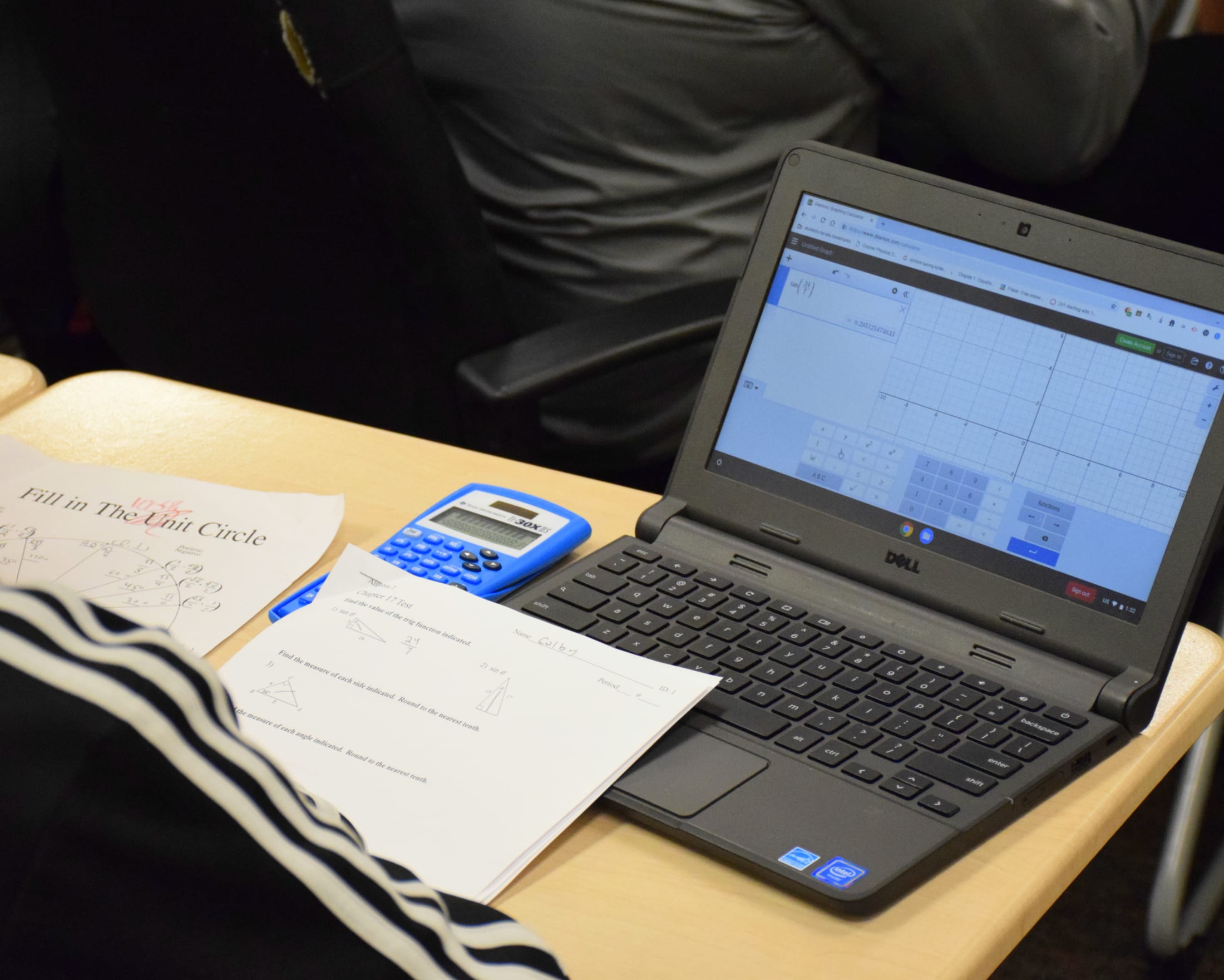 A laptop displaying a graph, a calculator, and a notebook on a desk, with a person's arm visible in the background.