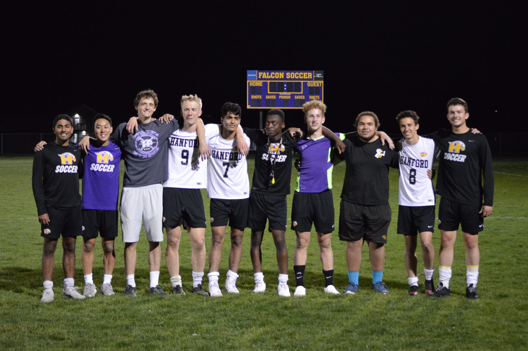 A group of young athletes, both male and female, standing together on a sports field at night, with a scoreboard visible in the background.