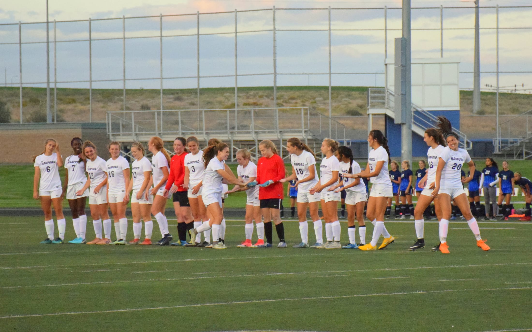 A group of female soccer players in white uniforms standing on a soccer field, with a stadium-like structure visible in the background.