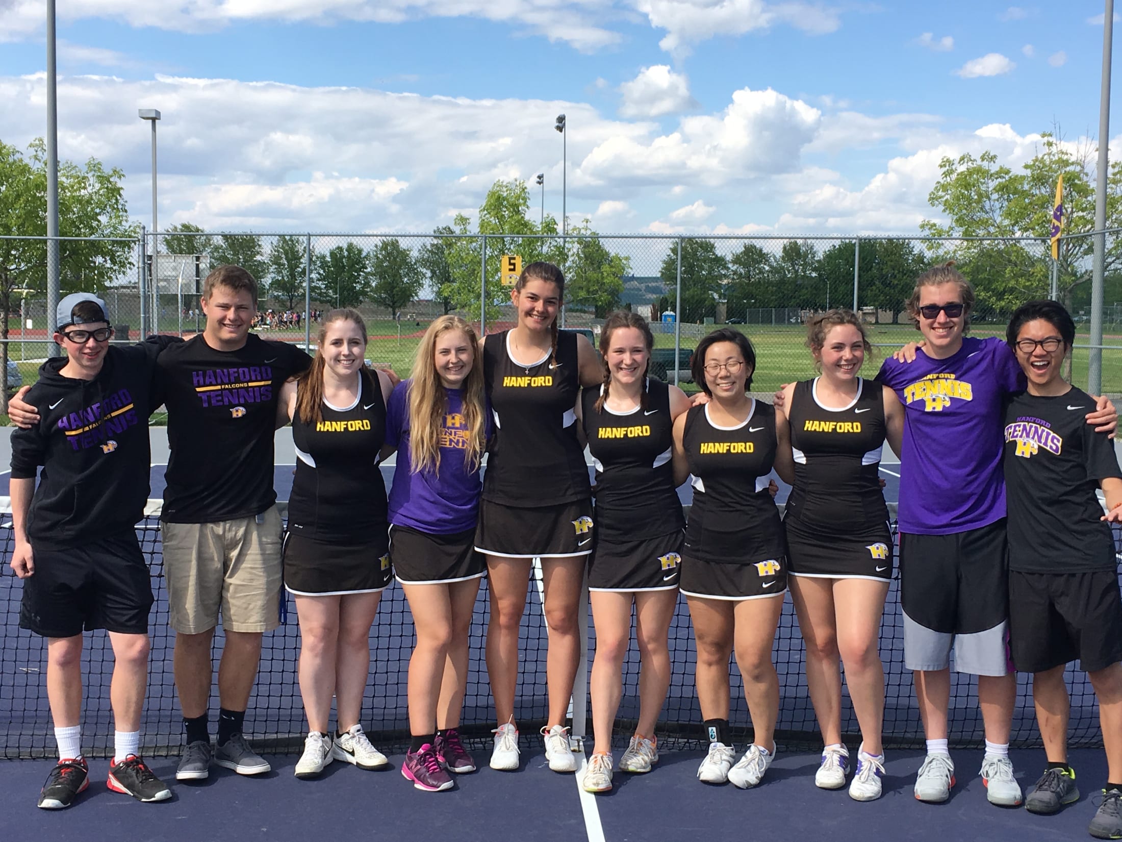 A group of young people, likely a sports team, posing together on a tennis court with a cloudy sky and trees in the background.