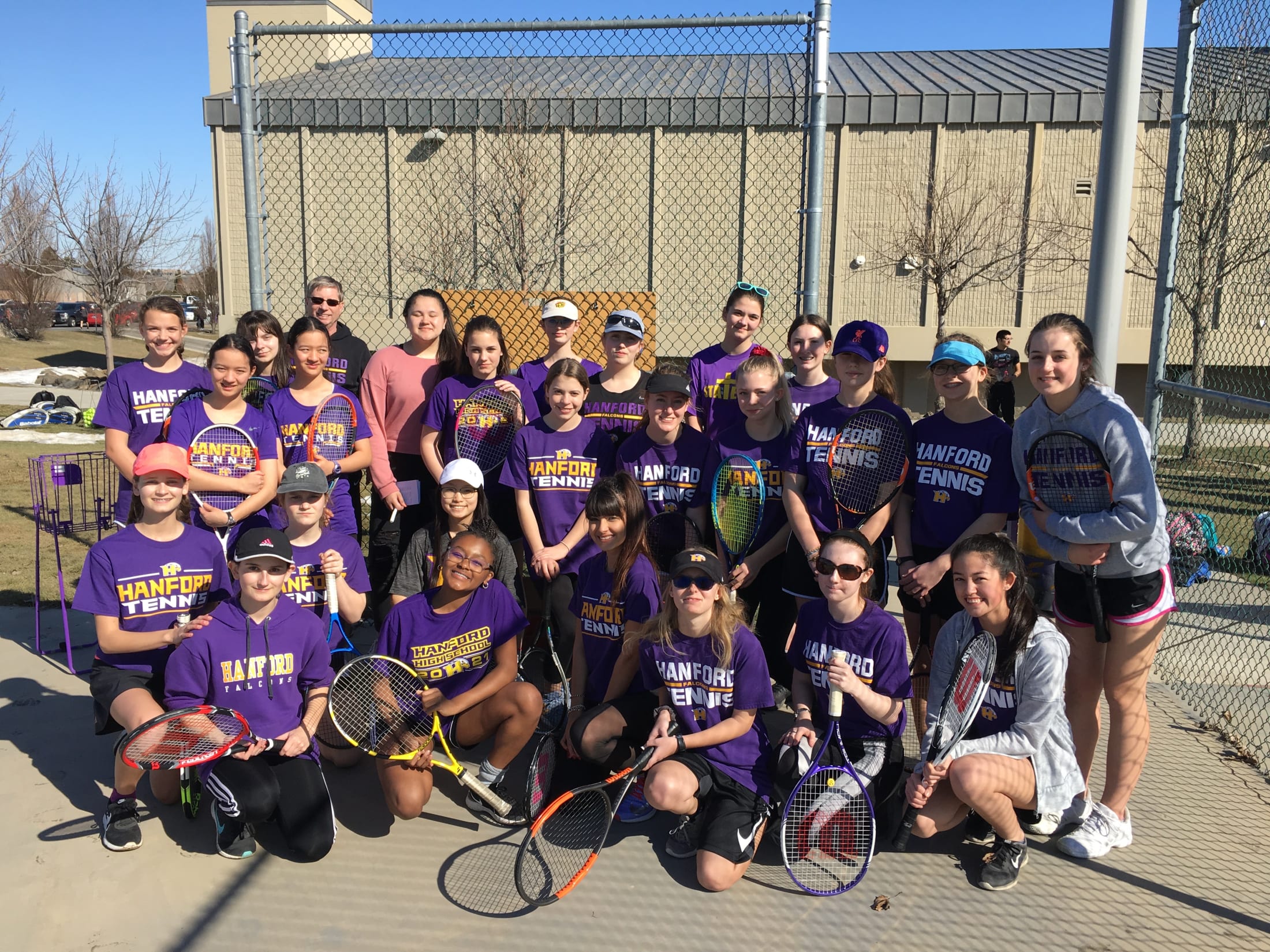 A group of people, likely a sports team, posing together outdoors on a tennis court with a chain-link fence and building in the background.