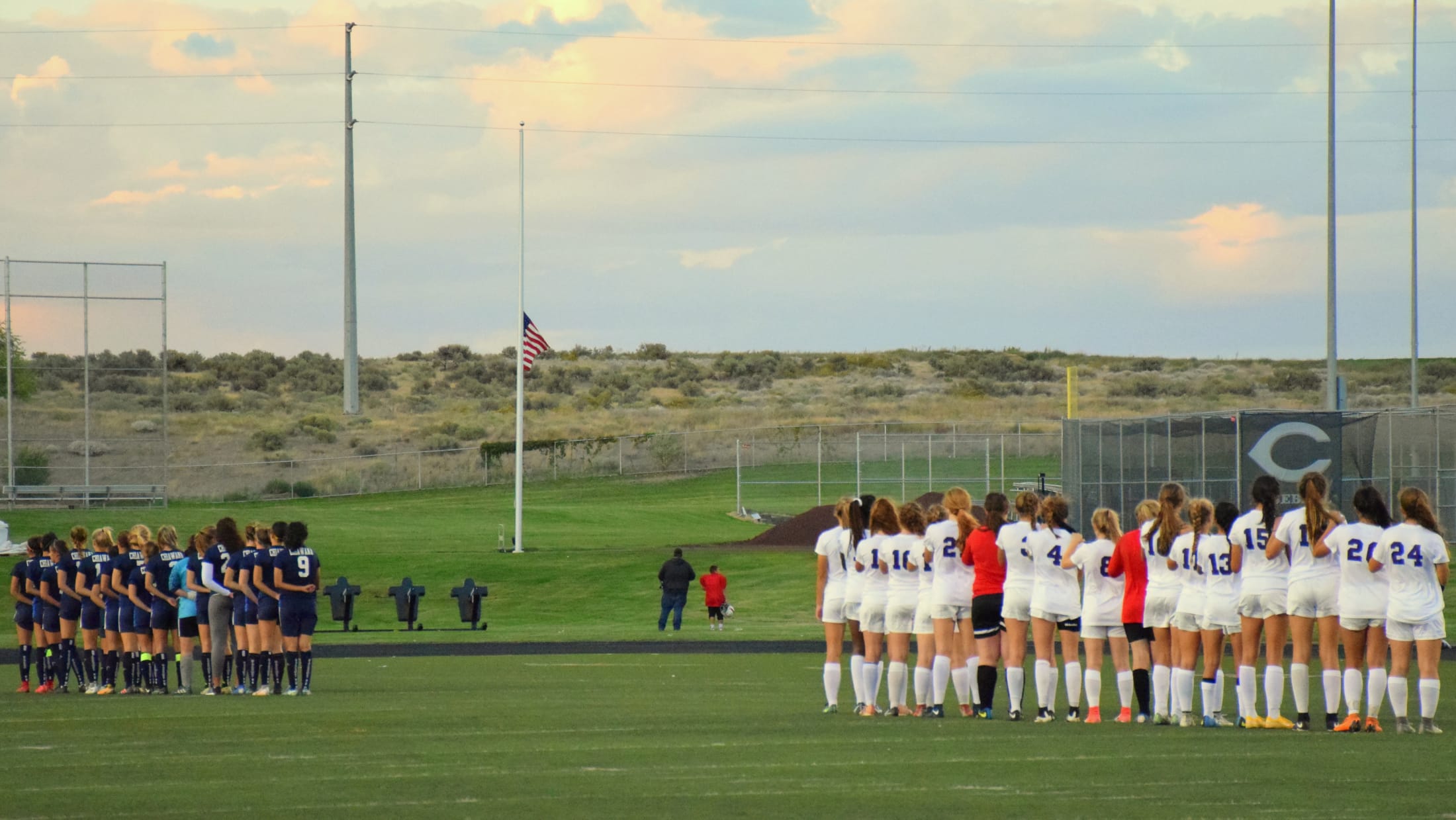 The image shows a soccer field with two teams lined up on the field, facing each other, with a flag pole and cloudy sky in the background.