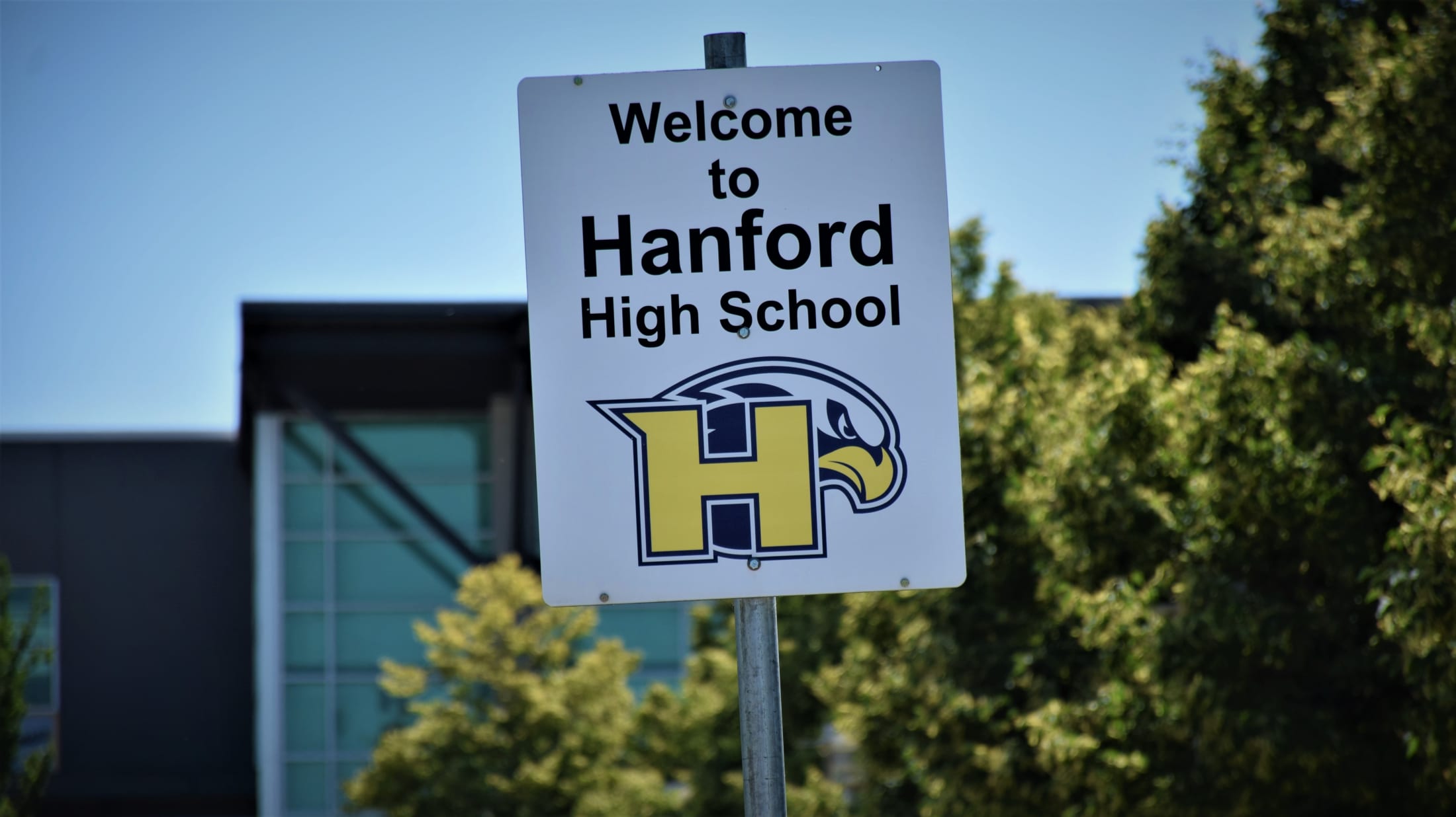 The image shows a welcome sign for Hanford High School, with the school's logo visible, set against a backdrop of trees and a clear blue sky.