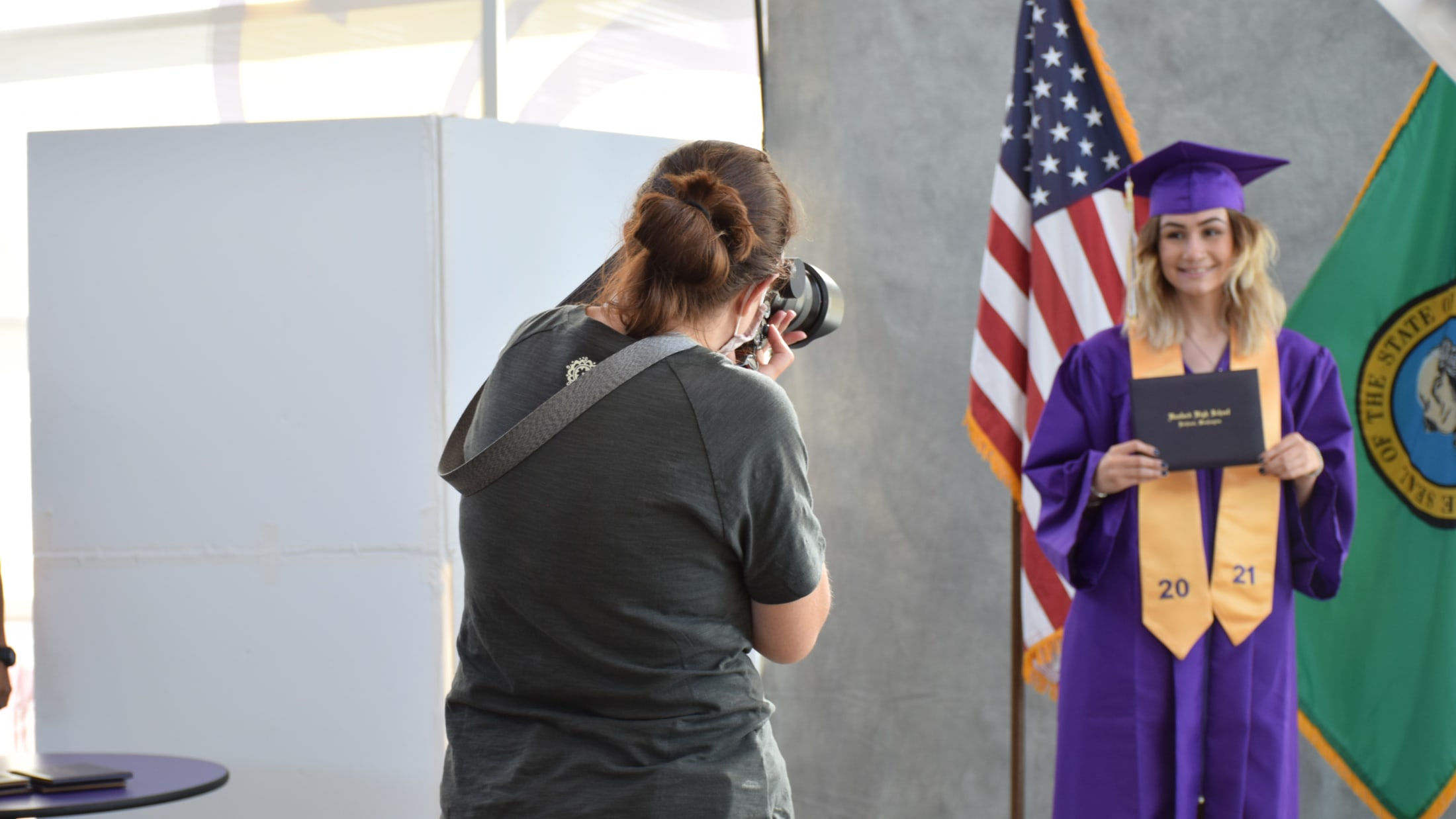 A young woman in a purple graduation gown stands in front of an American flag, while another person stands in the foreground.