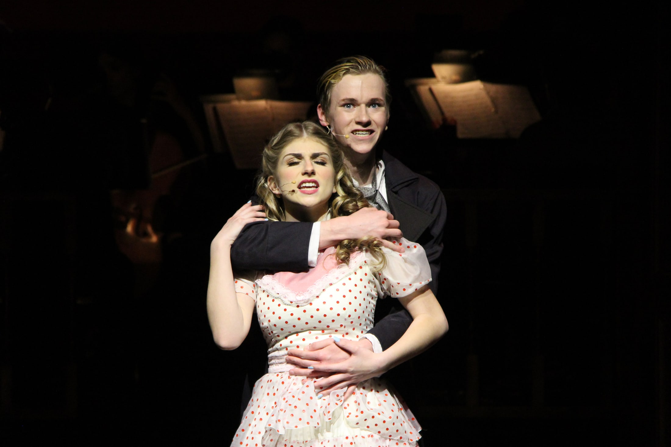 The image depicts a man and a woman in an intimate embrace, with the woman wearing a polka dot dress and the man wearing a dark suit. The background appears to be a dimly lit setting, with what looks like lamps or candles providing the only illumination.