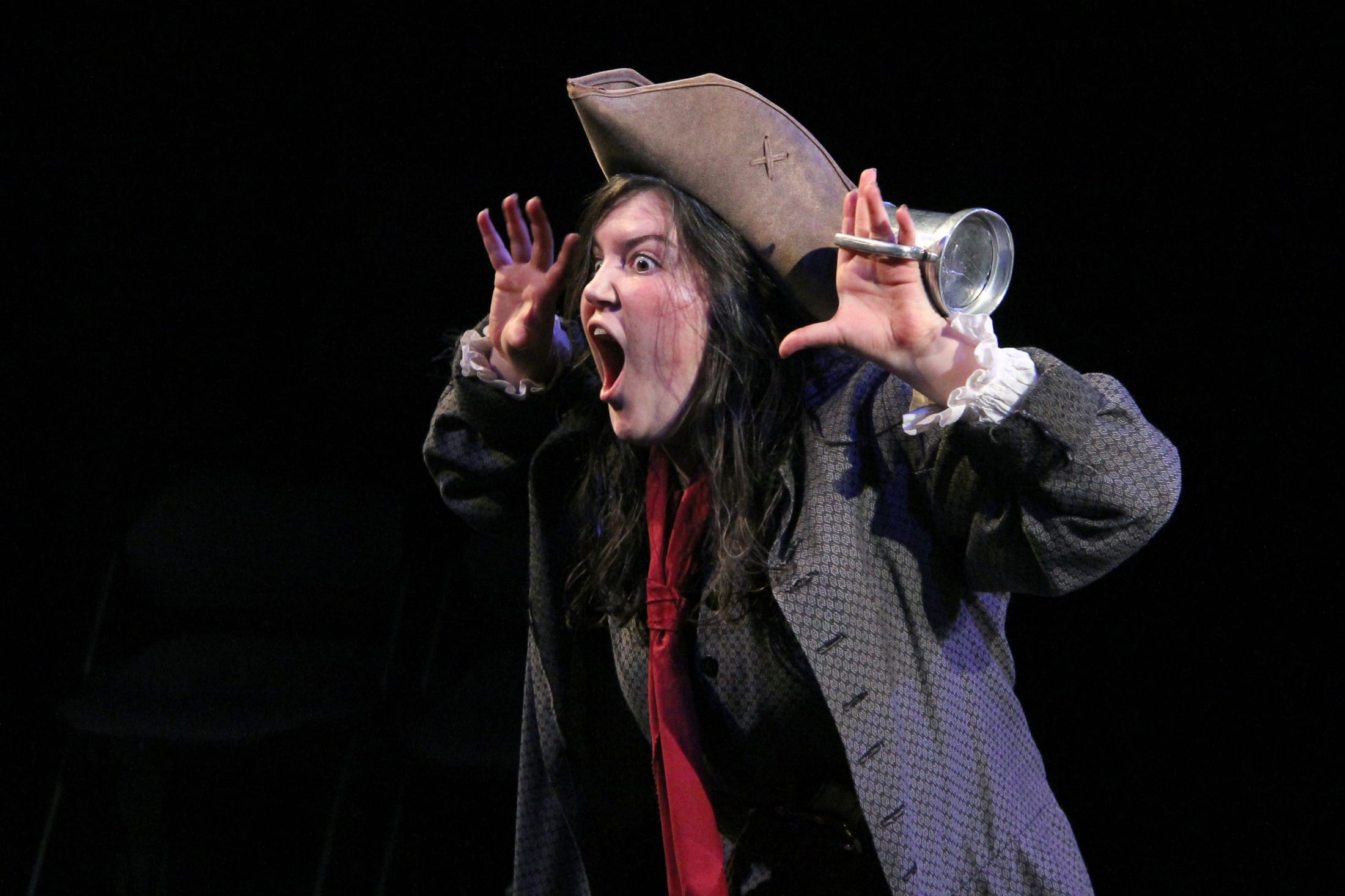 A person in a pirate-like costume with a tricorn hat and a red scarf is gesturing dramatically on a dark stage.