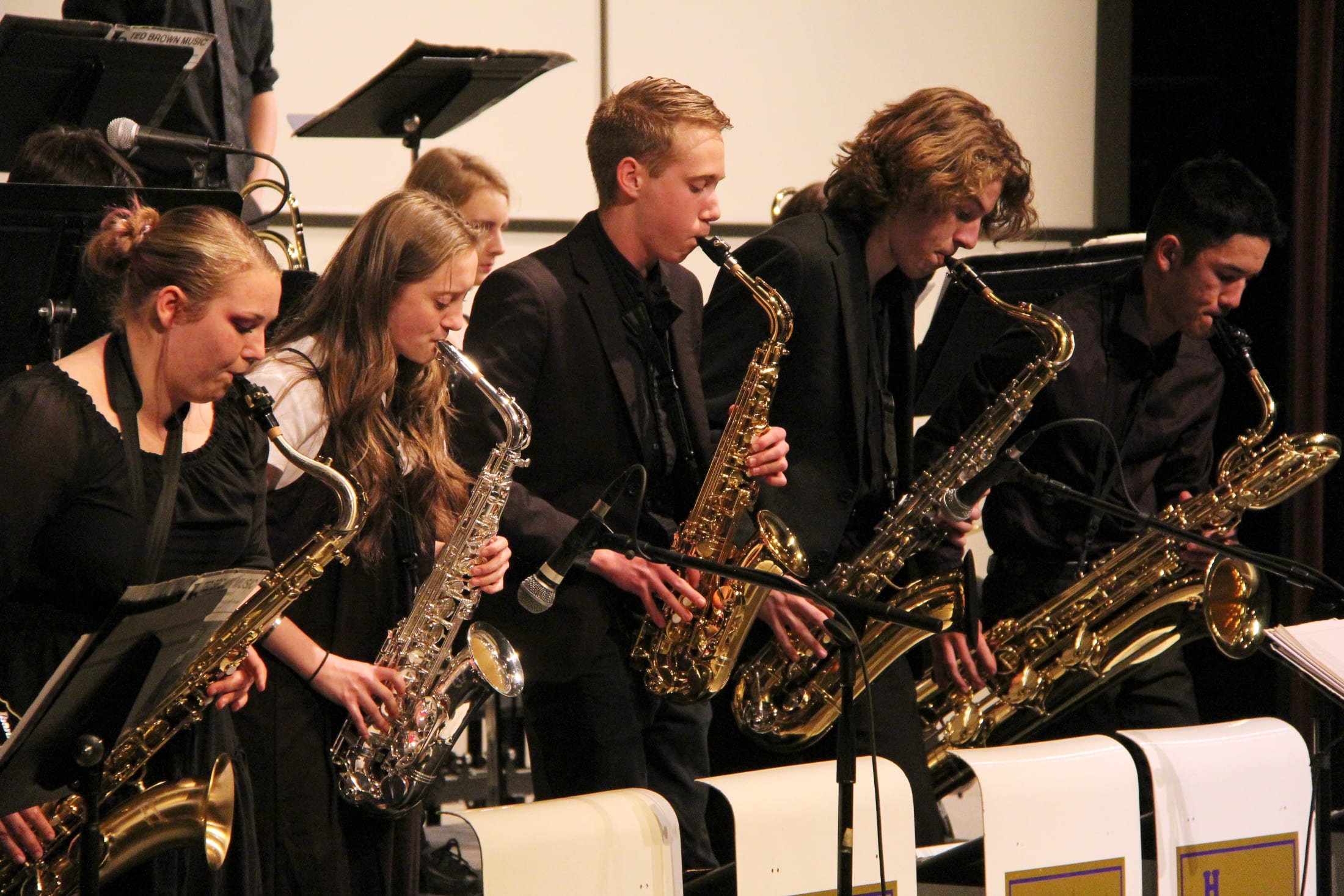 A group of musicians, dressed in black, are playing saxophones on a stage with musical equipment and chairs in the background.