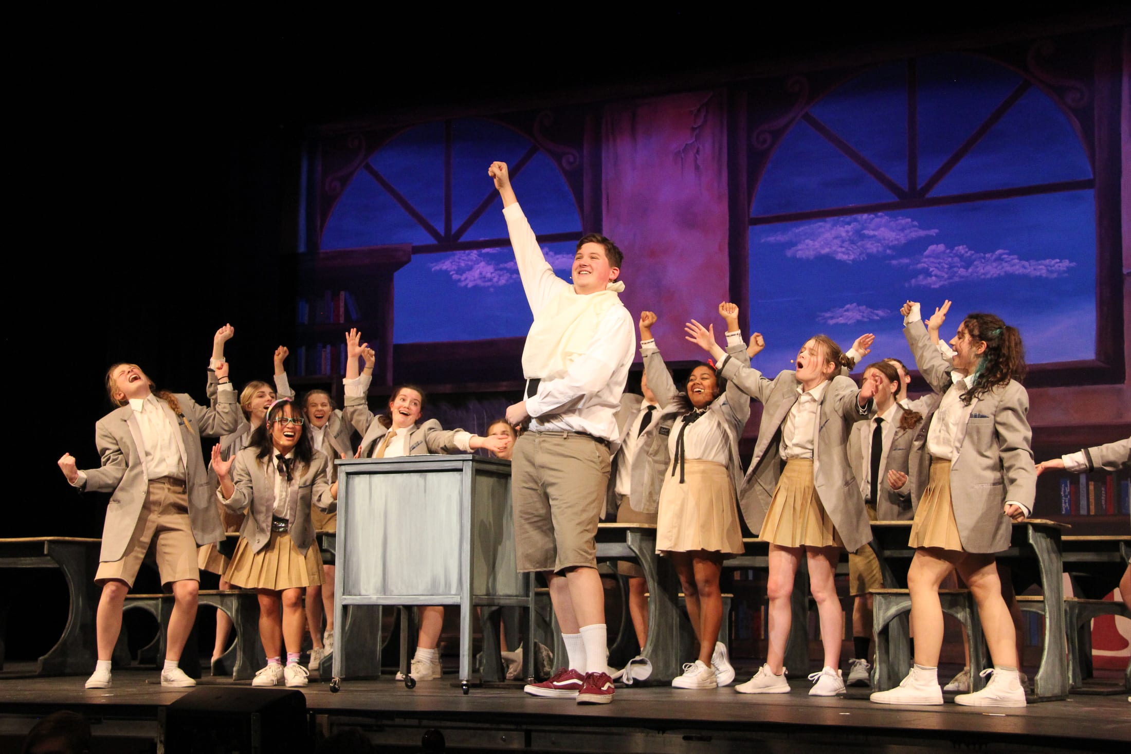 A group of young performers in uniform stand on a stage with a dramatic backdrop of a night sky, their arms raised in a unified gesture.