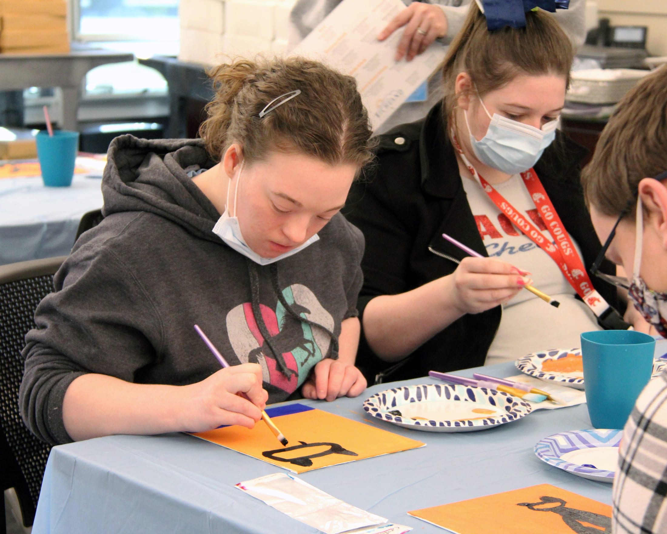 The image shows two young women wearing face masks and engaged in an arts and crafts activity, with various art supplies and materials visible on the table in front of them.