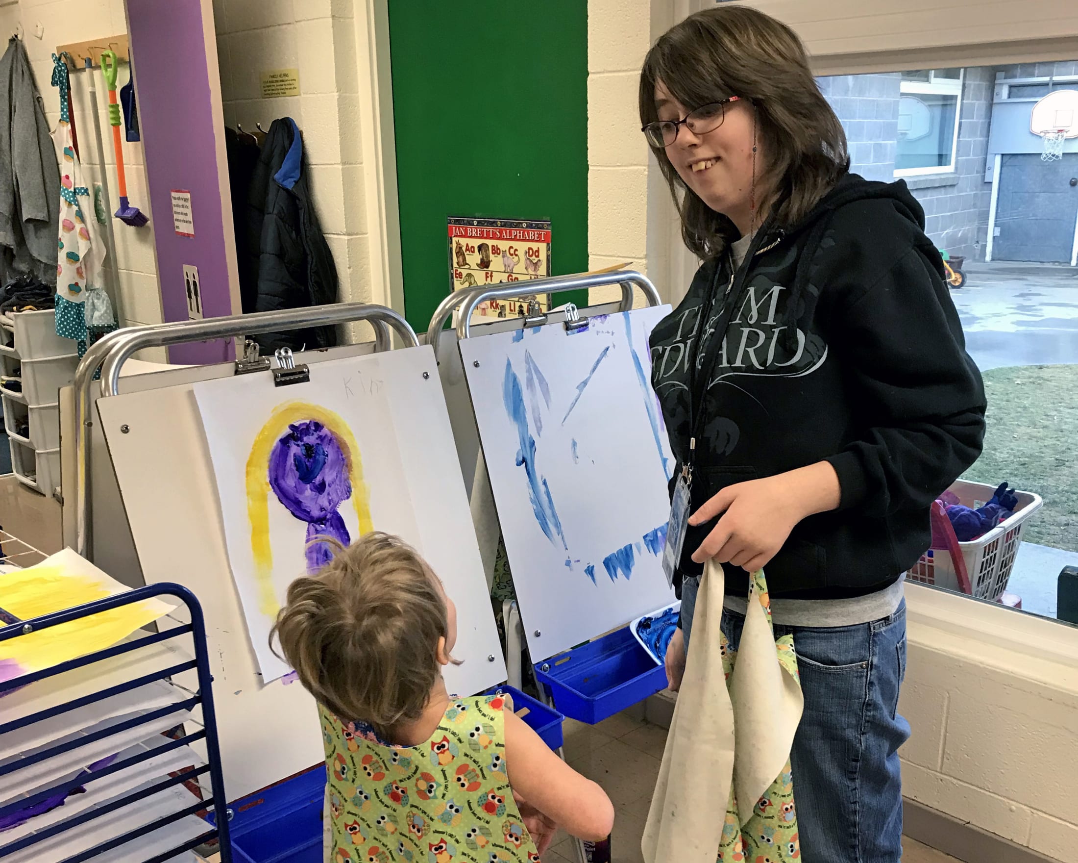 A young girl is standing in front of an easel, painting on a canvas, while an adult woman stands nearby observing her work.