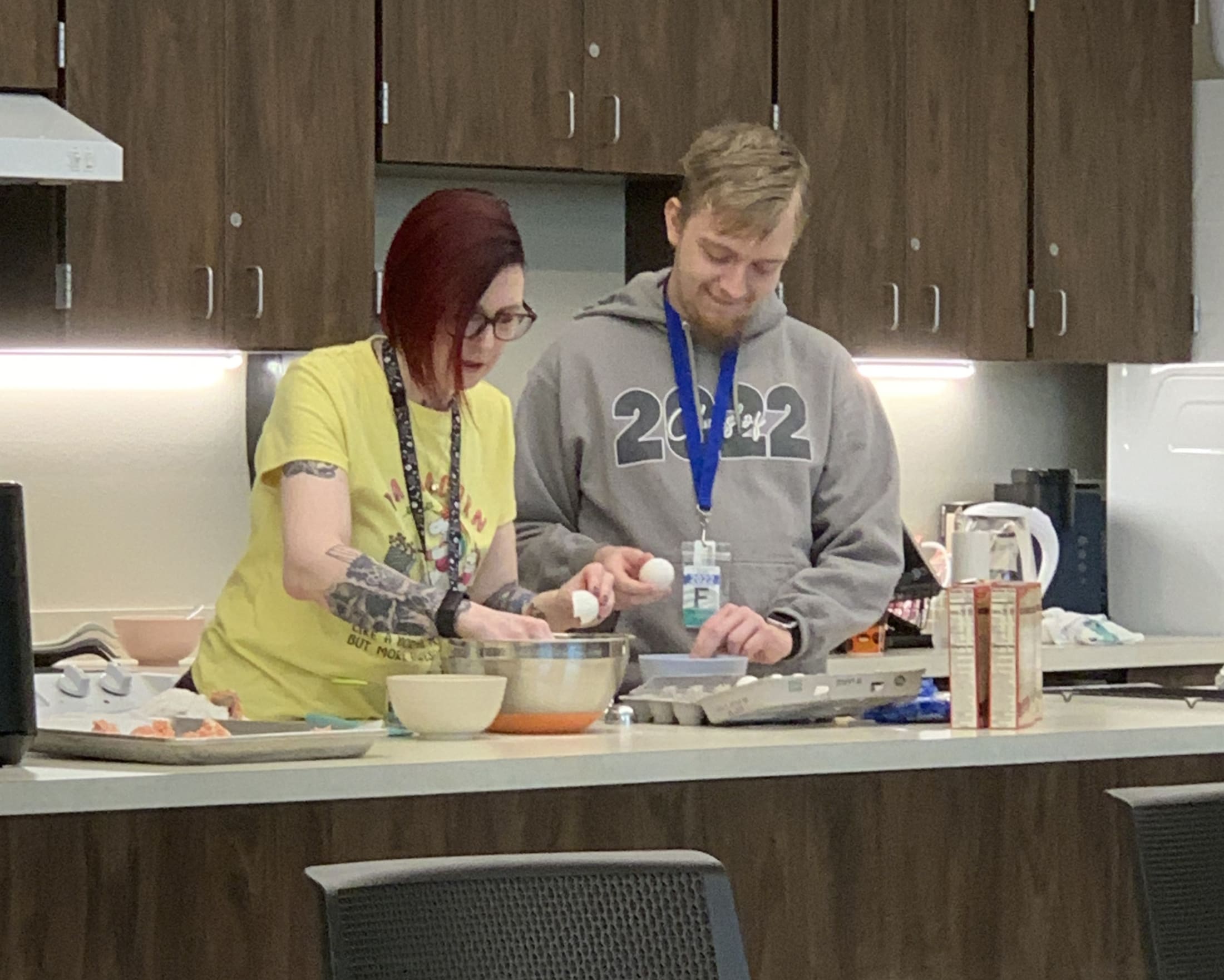 Two people, a woman wearing a yellow shirt and a man wearing a gray sweatshirt, are standing in a kitchen and preparing food together.