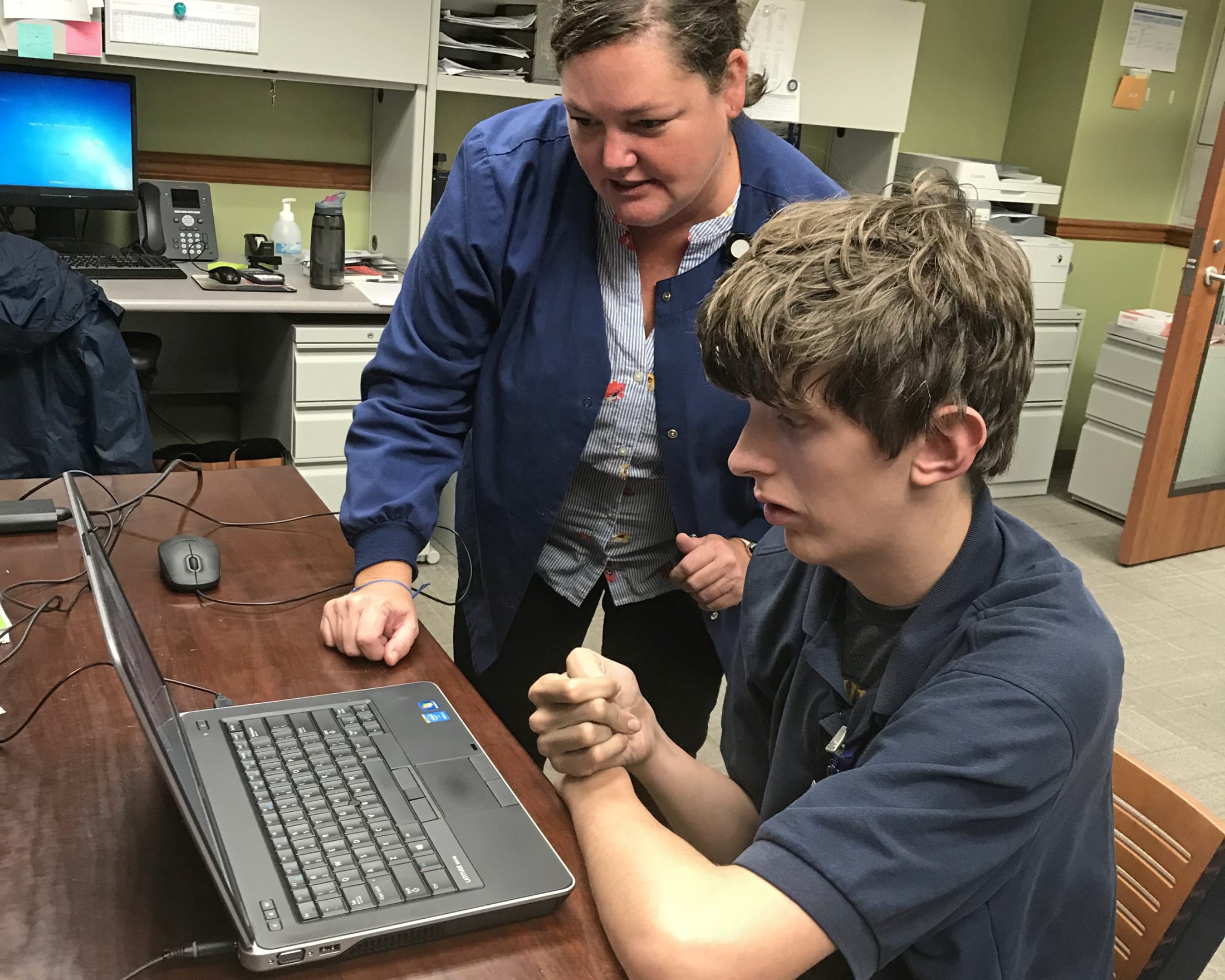 The image shows two individuals, an adult and a child, working together on a laptop computer in what appears to be an office or classroom setting.