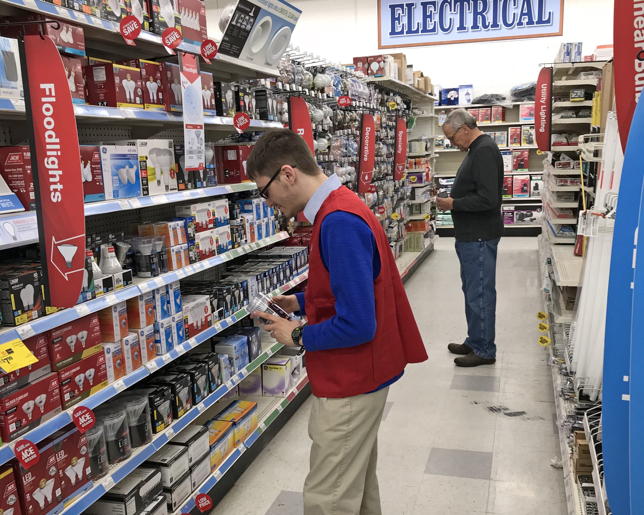 The image shows a man in a red jacket browsing the shelves of an electrical supply store, while another man stands in the background observing the store's offerings.
