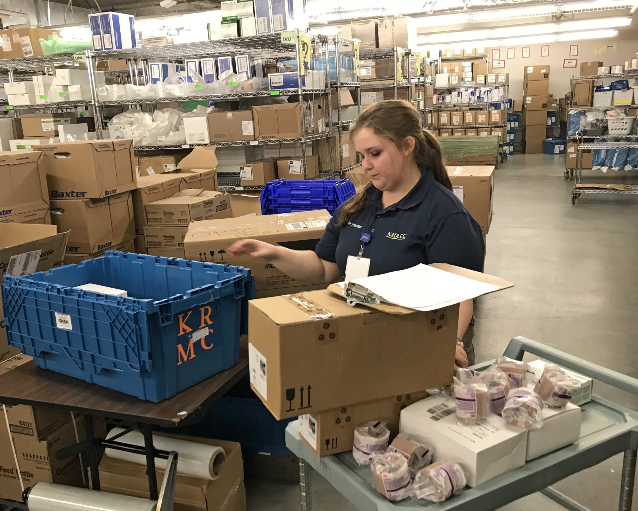 A woman in a blue uniform is working in a warehouse-like setting, surrounded by stacks of boxes and crates.