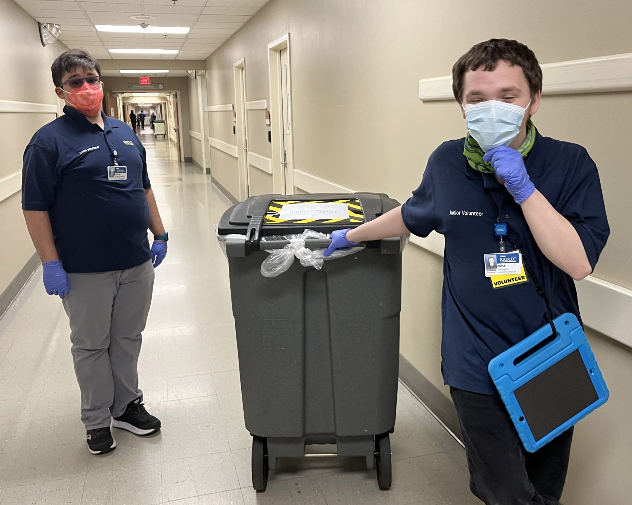 Two hospital staff members in uniform and masks are transporting a medical cart down a hospital hallway.