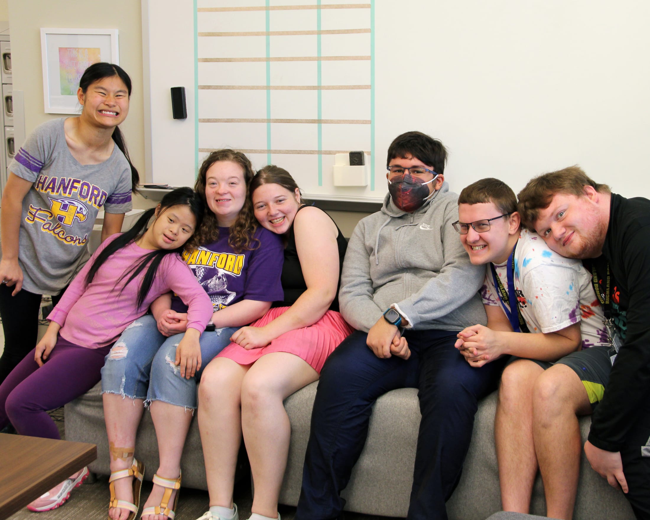 A group of smiling young people sitting together in a room with a white wall and windows in the background.