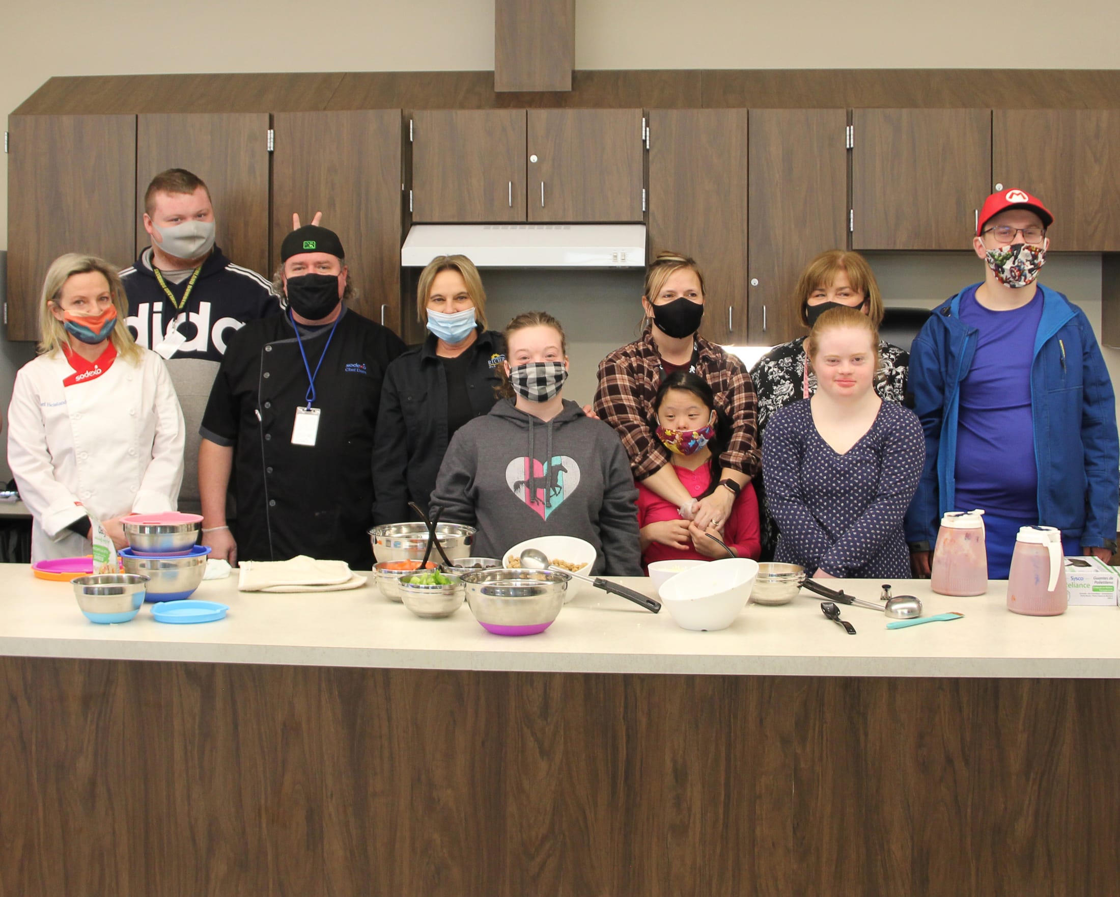 A group of people wearing masks are gathered around a kitchen counter, with various cooking supplies and utensils visible in the foreground.