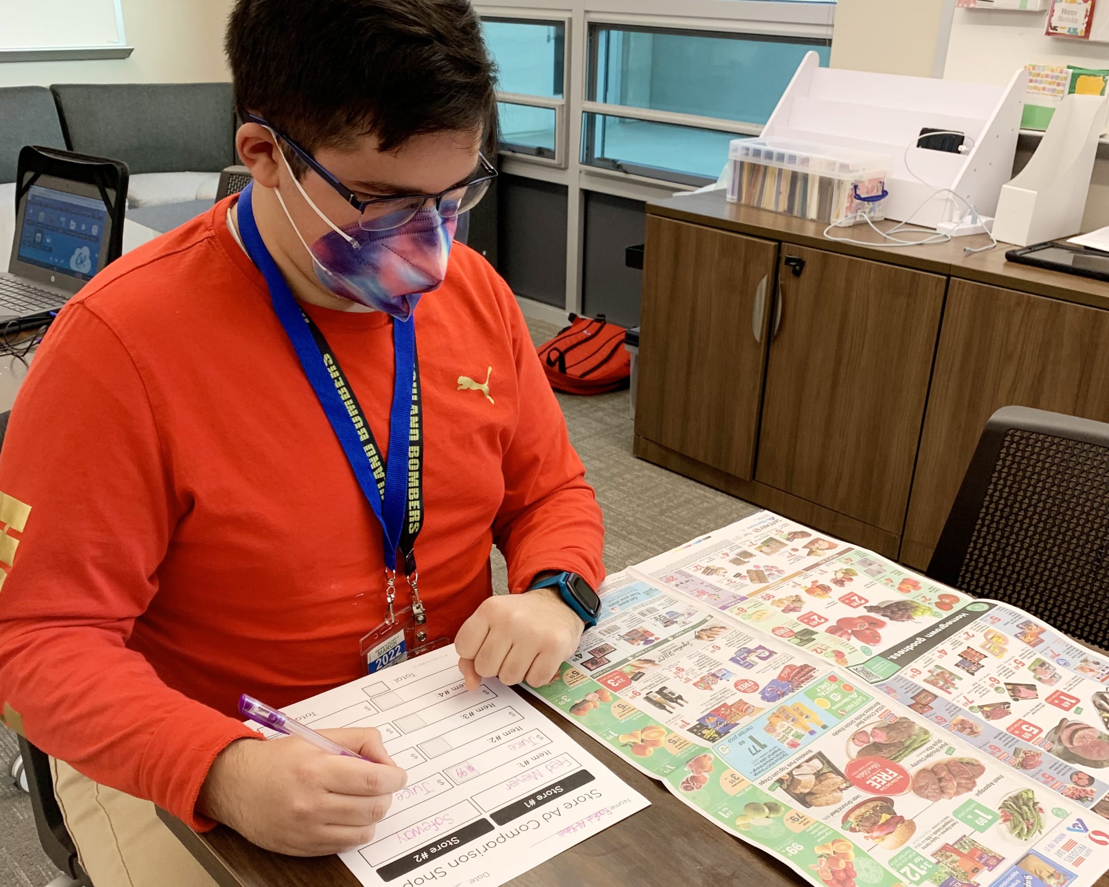 A person wearing a red shirt and a face mask is sitting at a desk, surrounded by various items and documents.