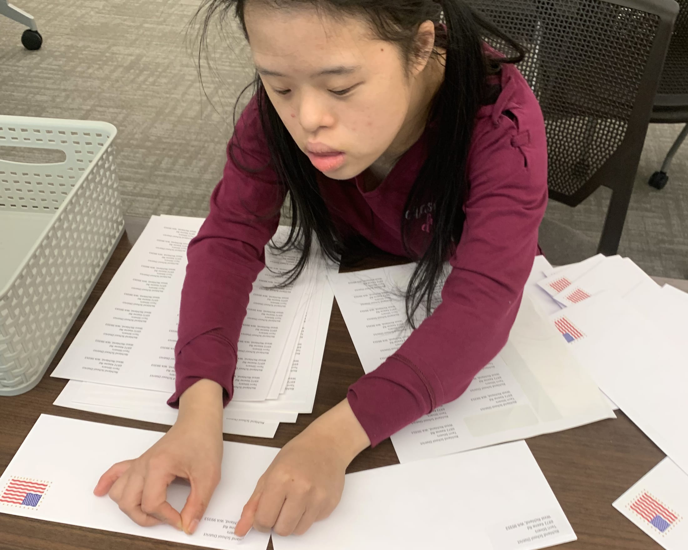 A young woman in a burgundy sweater is intently focused on reviewing documents spread out on a desk in an office setting.