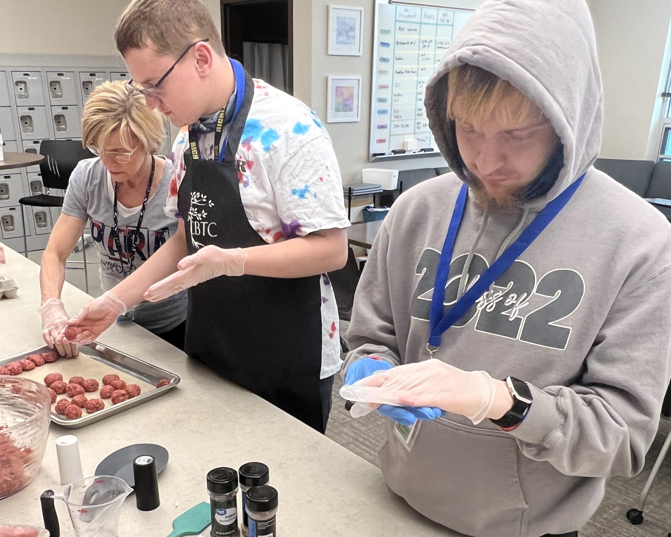 Two individuals, one wearing a hoodie and the other an apron, are working together to prepare food on a counter in what appears to be a kitchen or food preparation area.