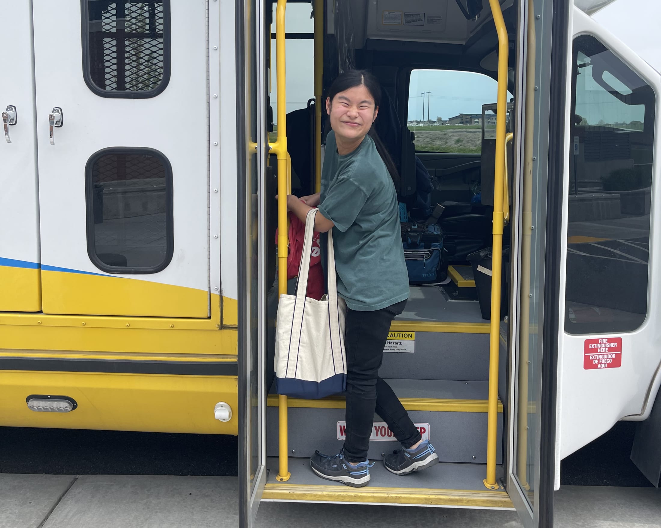 A person in a gray jacket is standing in the doorway of a yellow and white bus, holding a white bag.