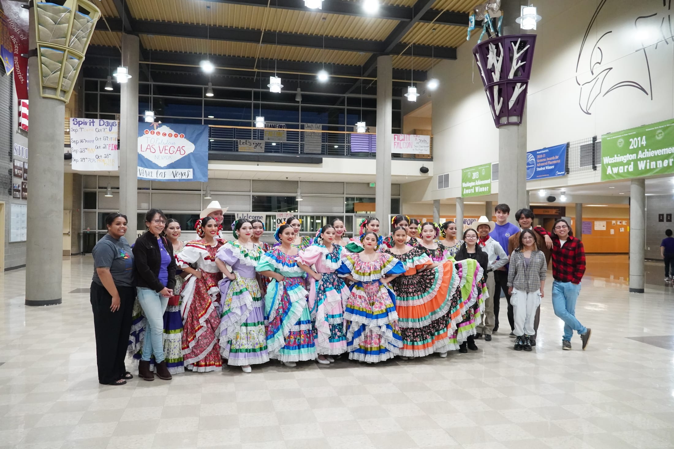 A group of people wearing colorful traditional dresses are gathered in a large indoor space with various signs and banners visible in the background.