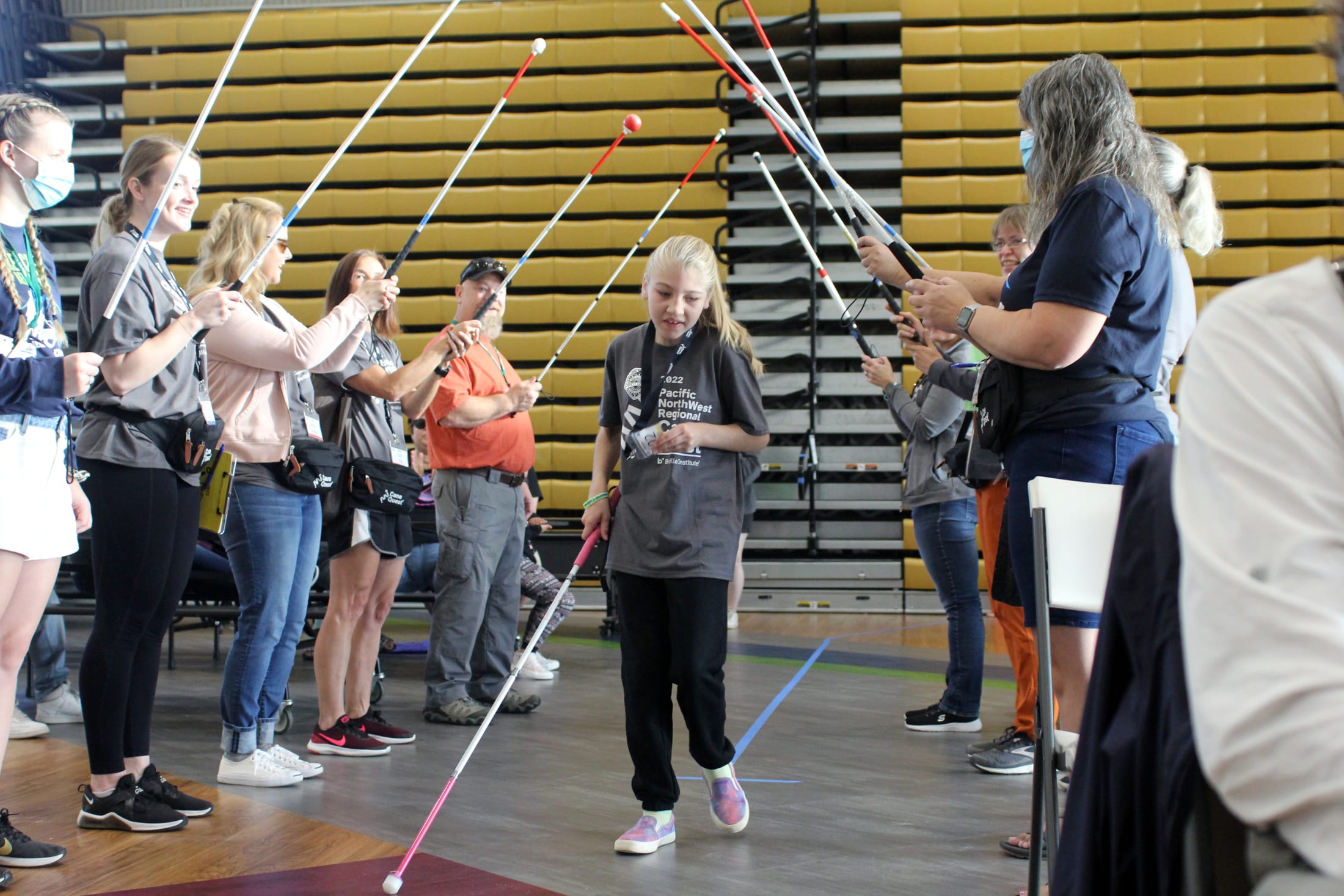 A group of people, some wearing casual clothing and others in more formal attire, are gathered in what appears to be a gymnasium or indoor sports facility, with yellow bleachers visible in the background.