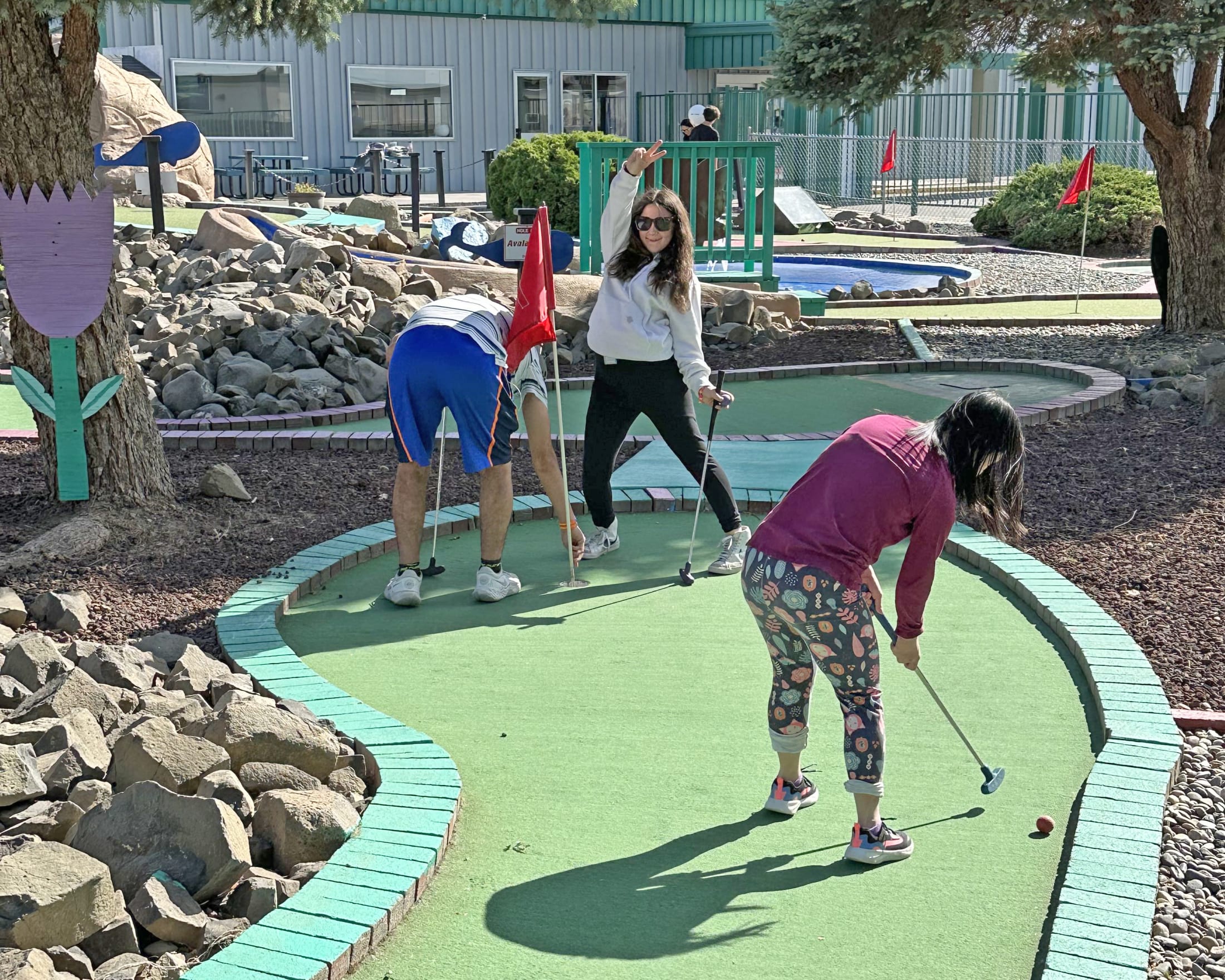 The image shows a group of people playing mini golf on a colorful, winding course surrounded by rocks, trees, and a playground in the background.