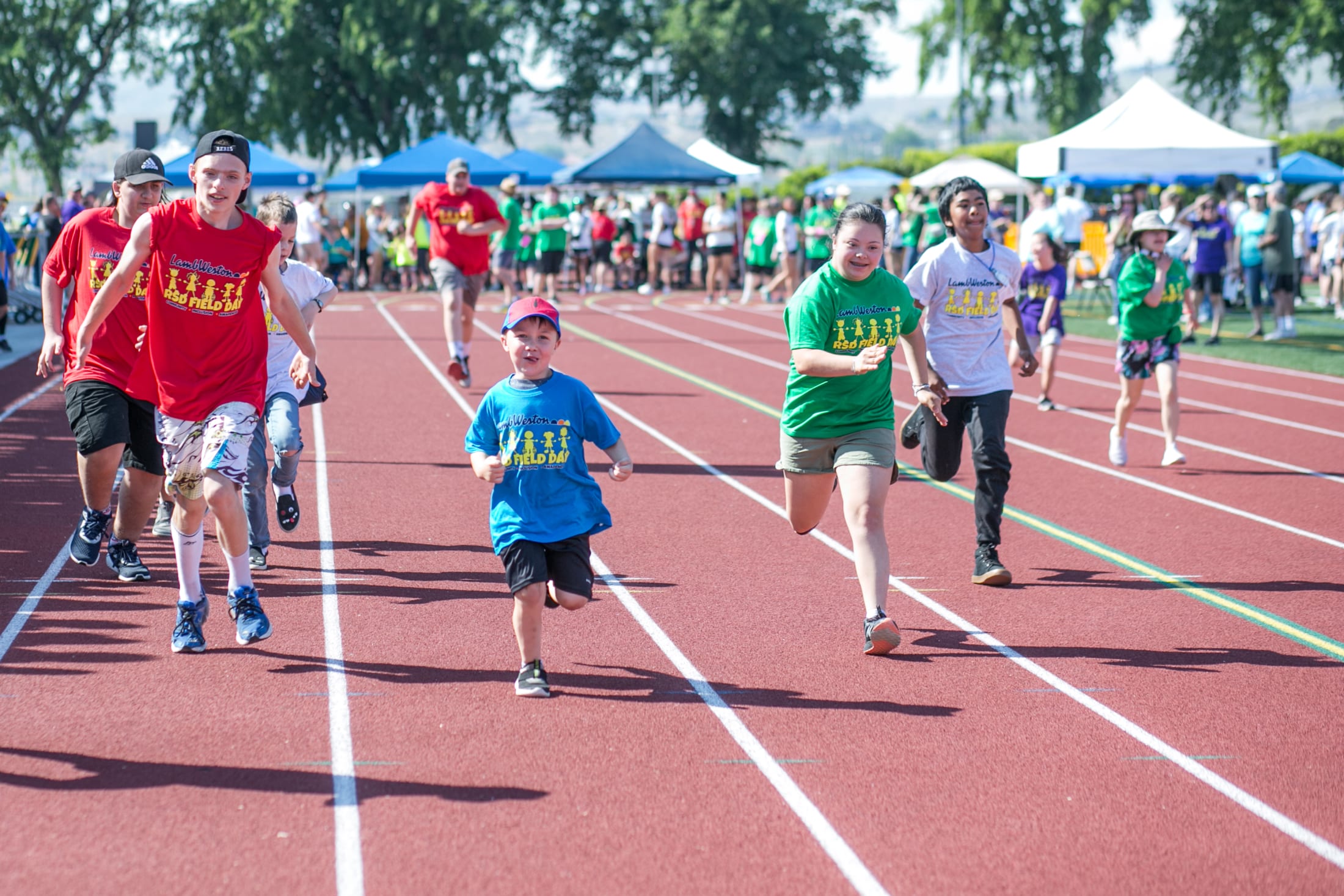 A group of people, some wearing colorful shirts, are walking on a running track surrounded by tents and trees in the background.