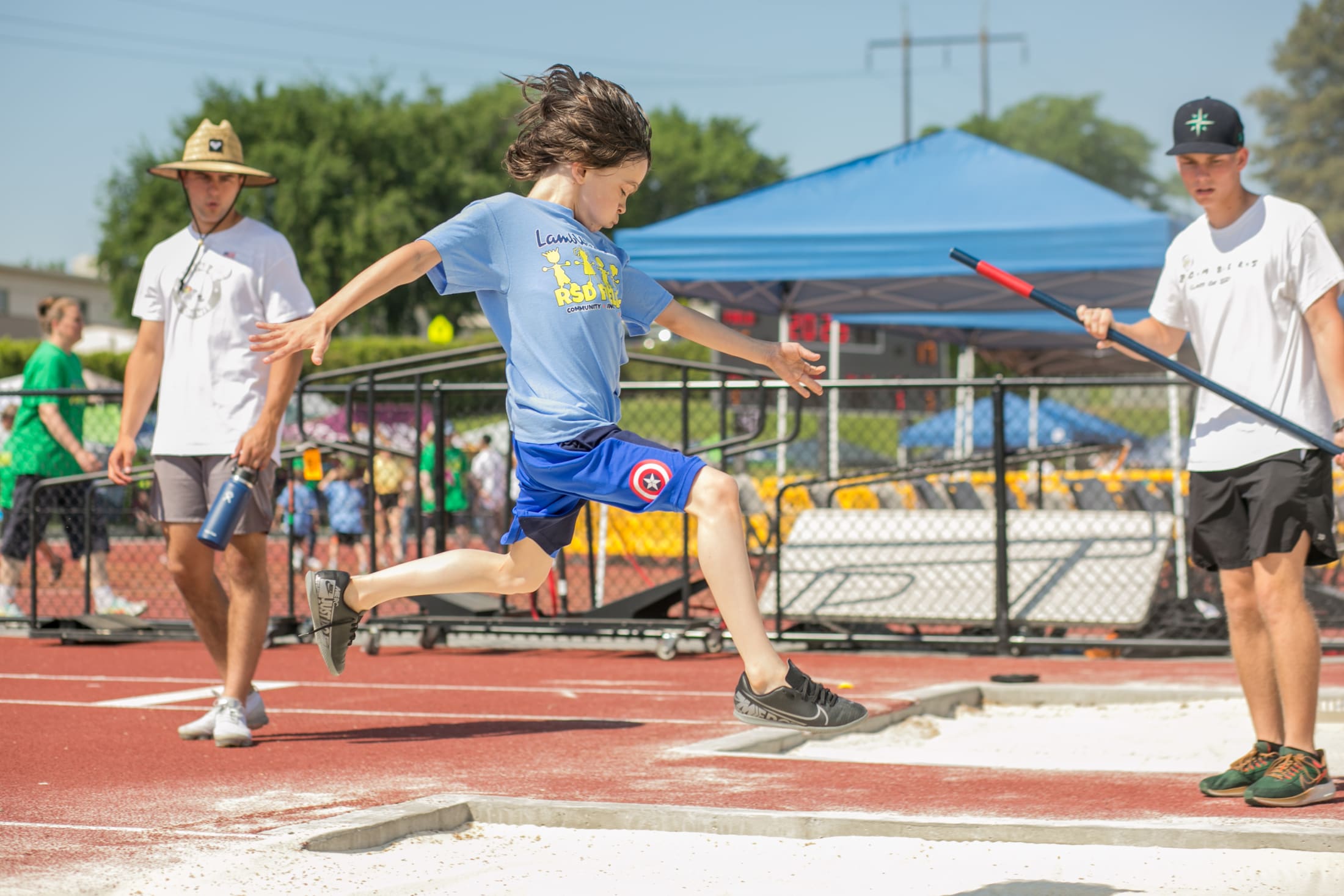 The image shows a group of people participating in a track and field event, with a person in the foreground preparing to jump on a red track surrounded by tents and other spectators in the background.
