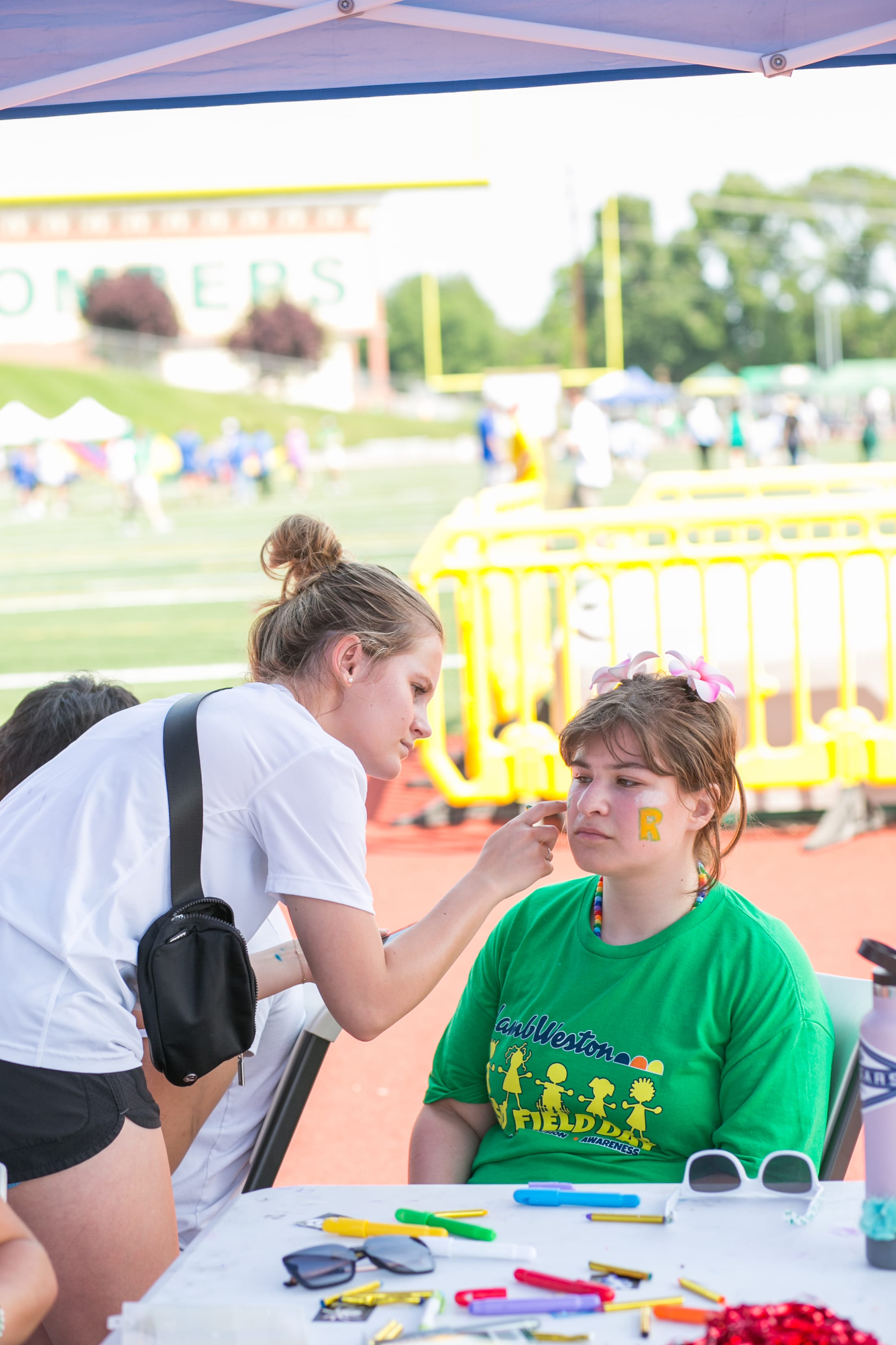 A woman in casual clothing is interacting with a young child wearing a green t-shirt, both seated at a table with various art supplies in an outdoor setting with a carnival or fair in the background.