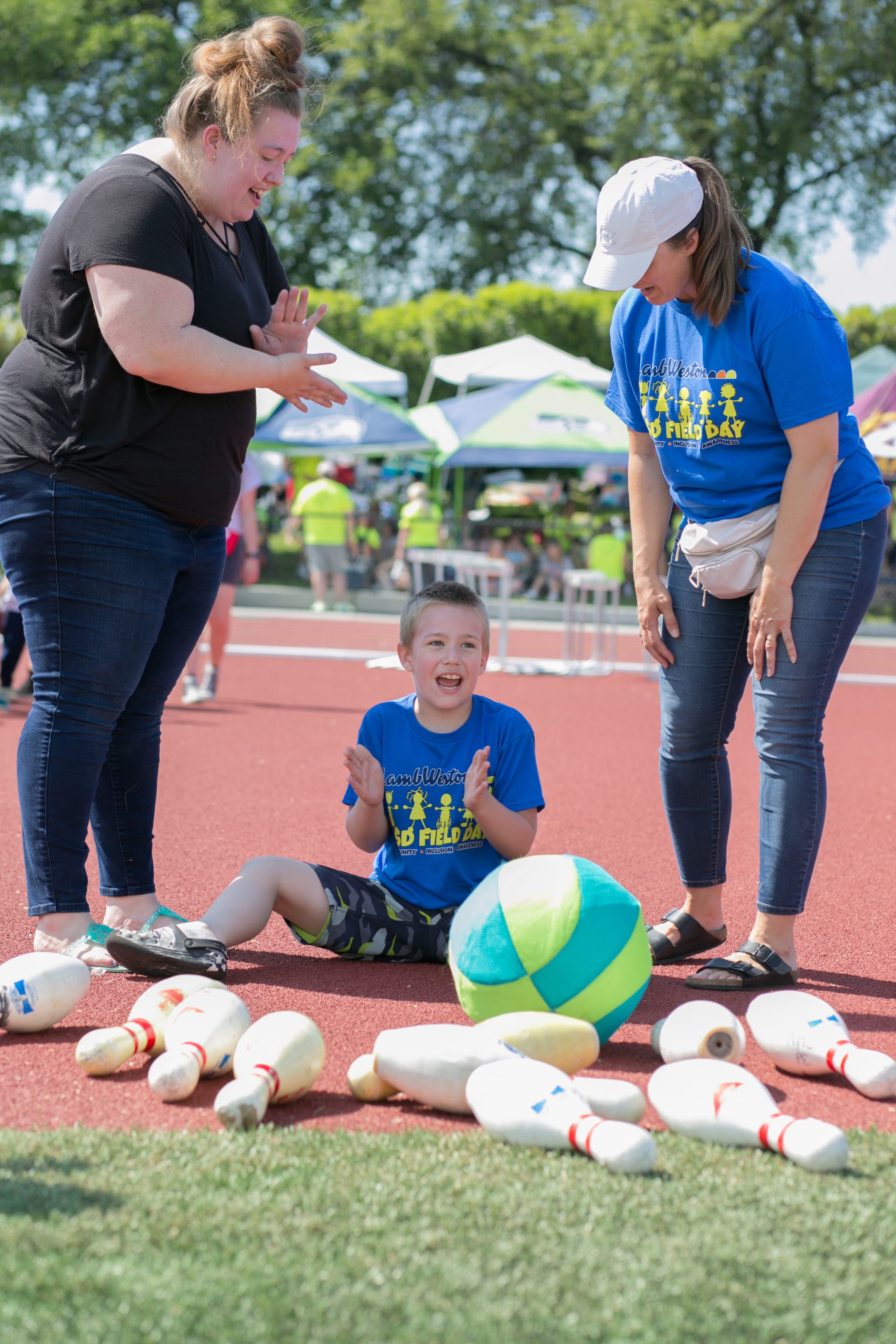The image shows a young child sitting on a rubber surface surrounded by various sports equipment, with two adults, one wearing a blue shirt and the other a black shirt, interacting with the child.