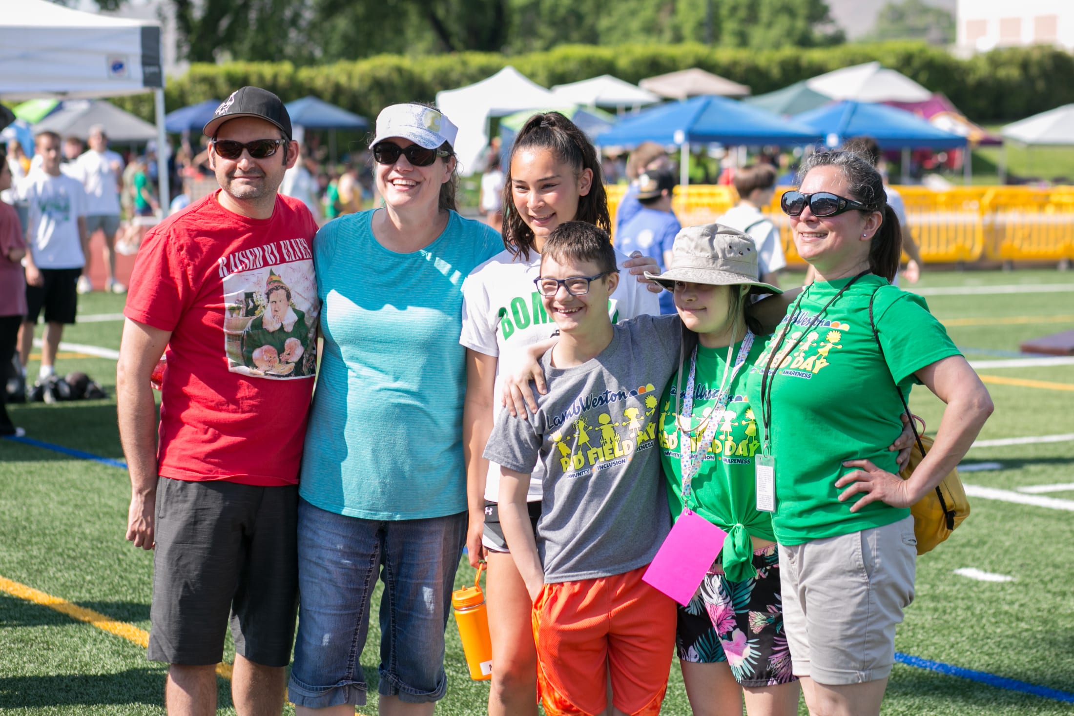 A group of people, including adults and children, are standing together in a park-like setting with tents and other outdoor activities visible in the background.