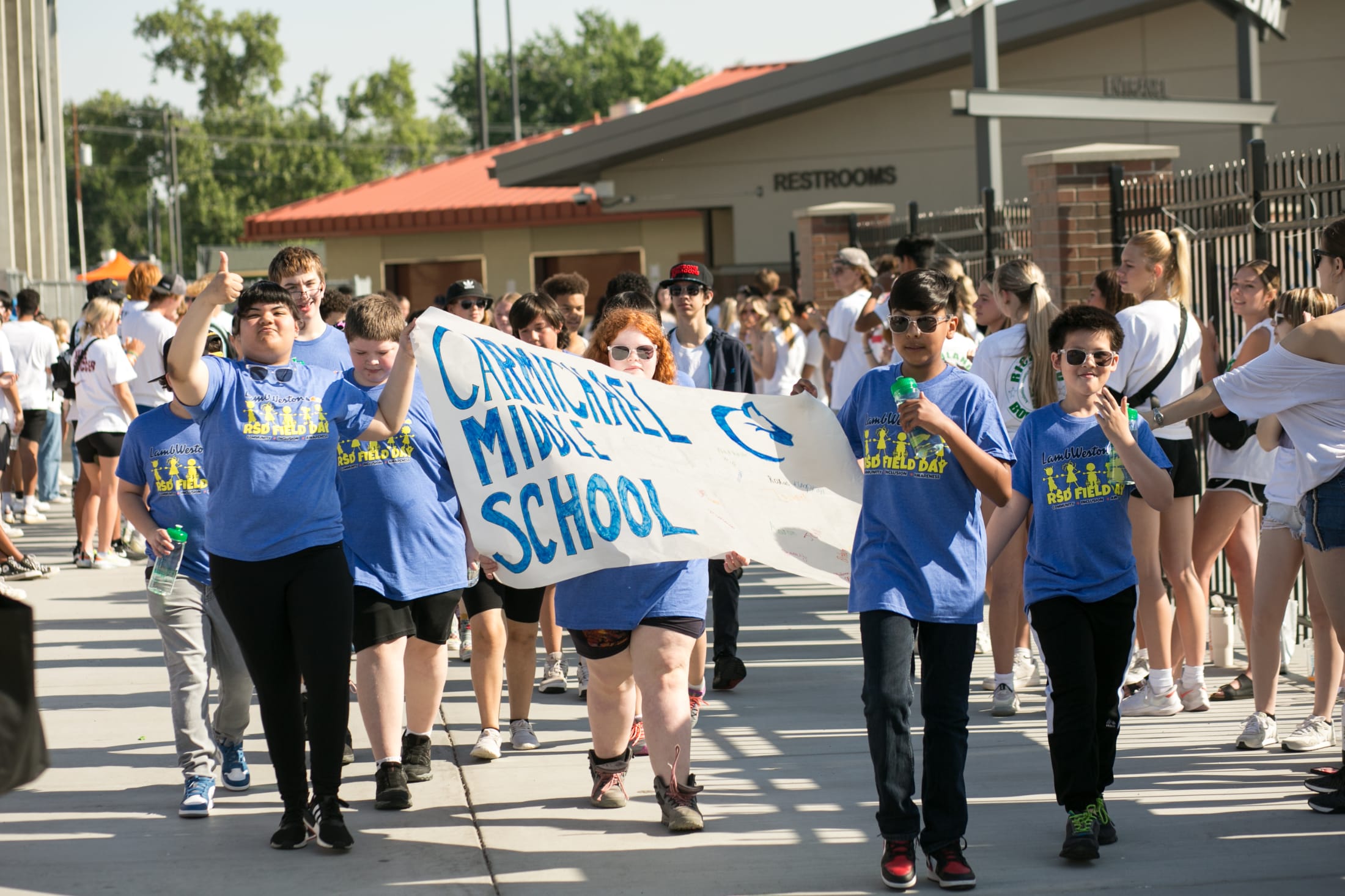 A group of people, mostly students, are walking together and holding a banner that says %22Camarillo High School%22 as they appear to be participating in some kind of event or demonstration in front of a building with a red roof.