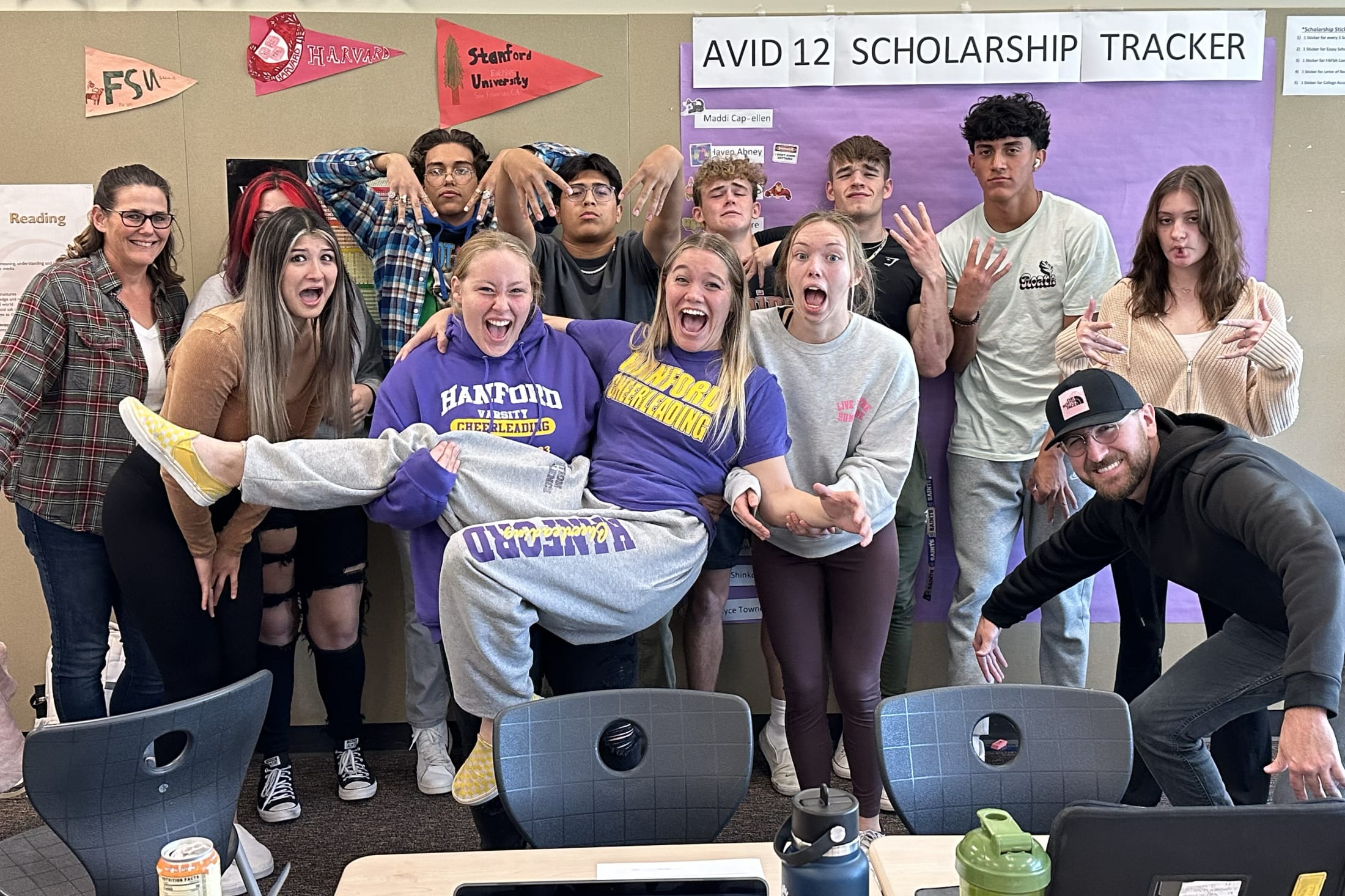 A group of enthusiastic students posing together in a classroom setting, with various educational posters and materials visible in the background.